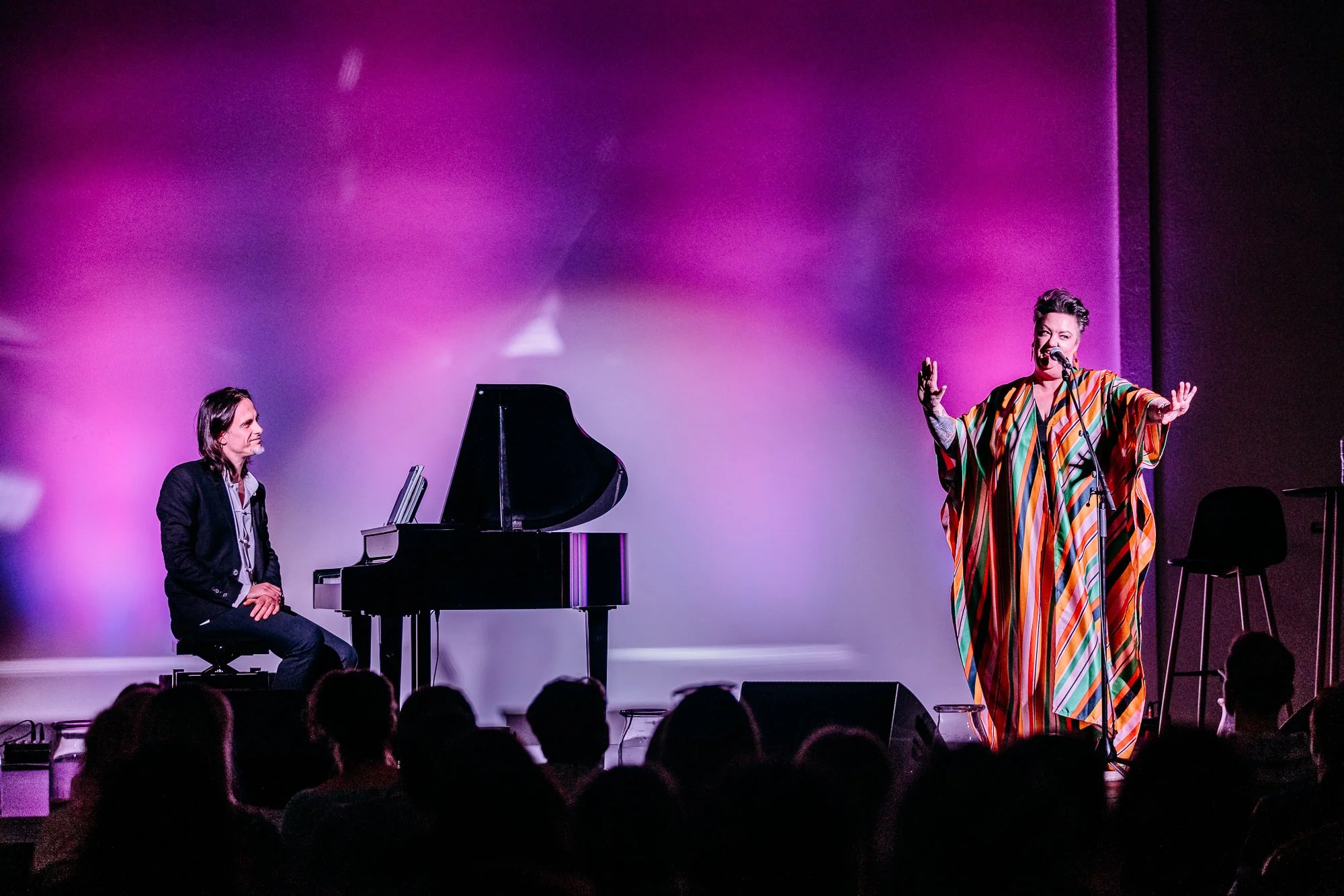 A woman singing into a microphone on stage, wearing a colorful striped dress, with a pianist sitting at a black grand piano nearby. The audience is visible in silhouette.