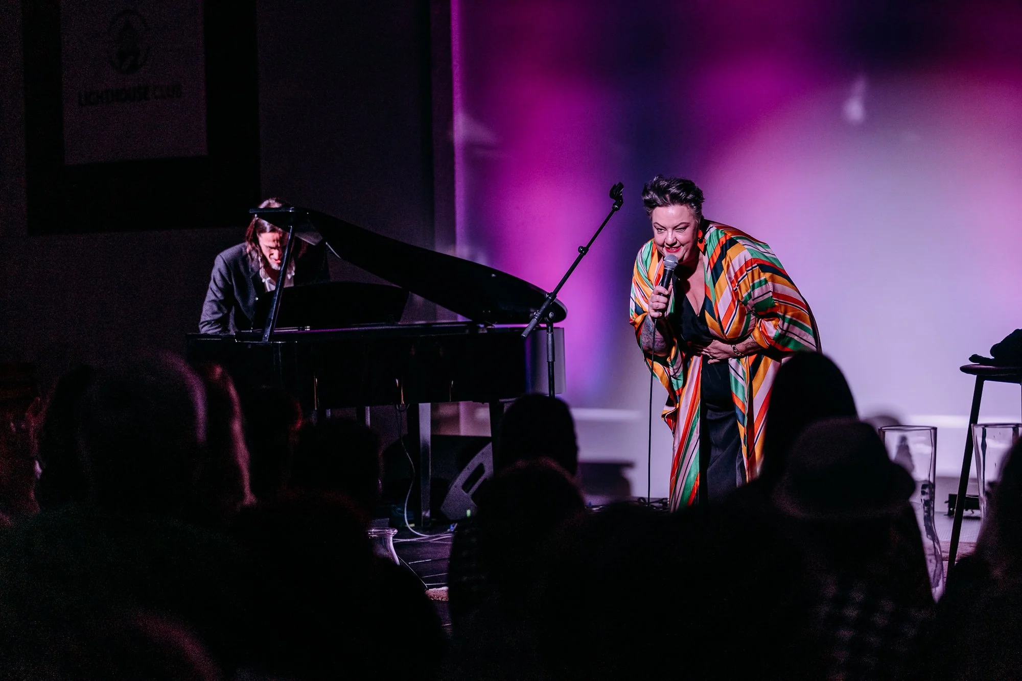 A woman singing into a microphone while bending forward on stage, with a pianist playing a grand piano beside her. The stage is lit with purple lighting, and an audience is seated in front.