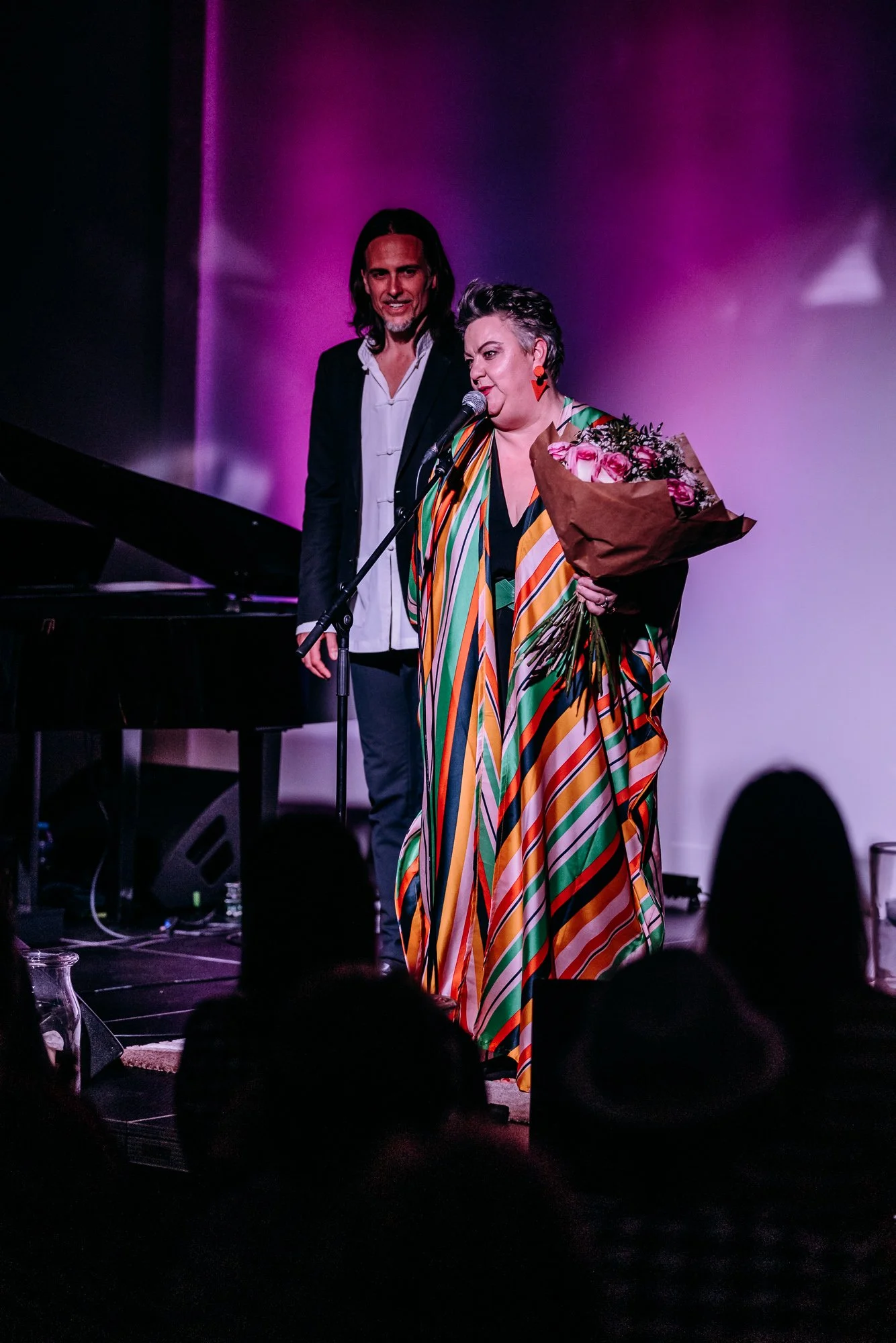 A woman with short, gray hair in colorful striped dress holding a bouquet of pink roses on stage with a man in a black suit behind her, in front of a purple-lit background, at a performance or event.