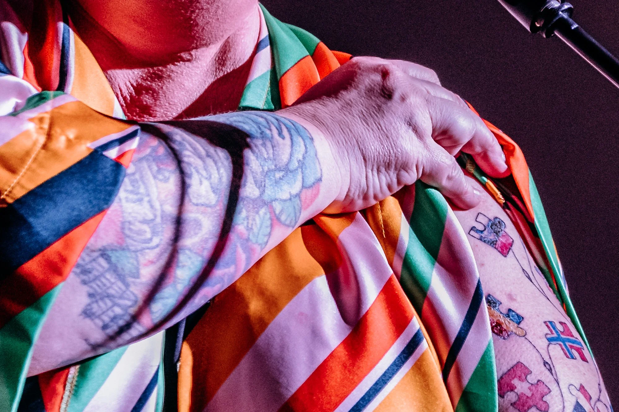Close-up of a person's hand with a tattoo, resting on their chest, wearing colorful clothing with various patterns.