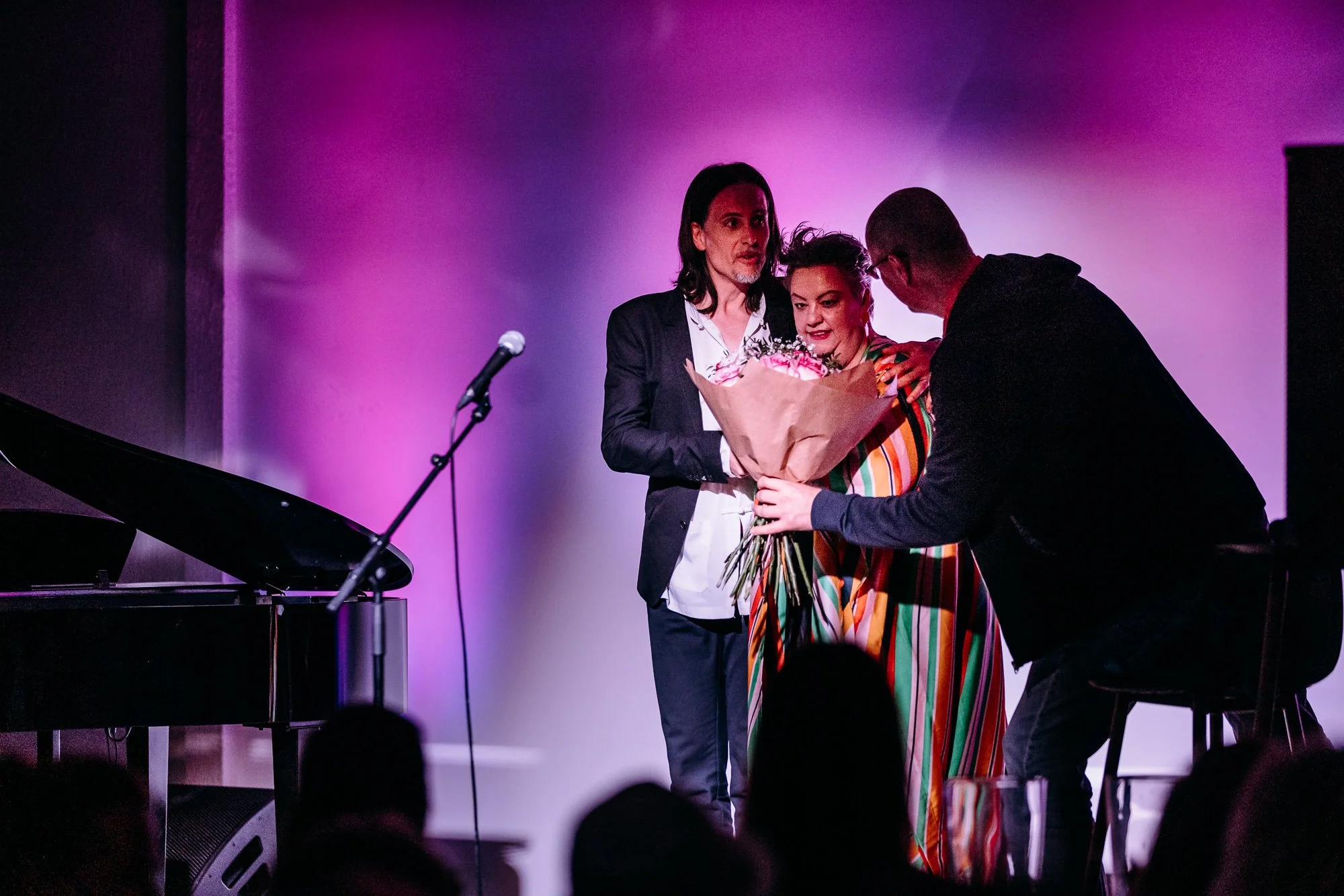 Three people on stage, one man handing a bouquet of flowers to a woman in colorful striped dress, with purple lighting background, microphone and piano nearby.