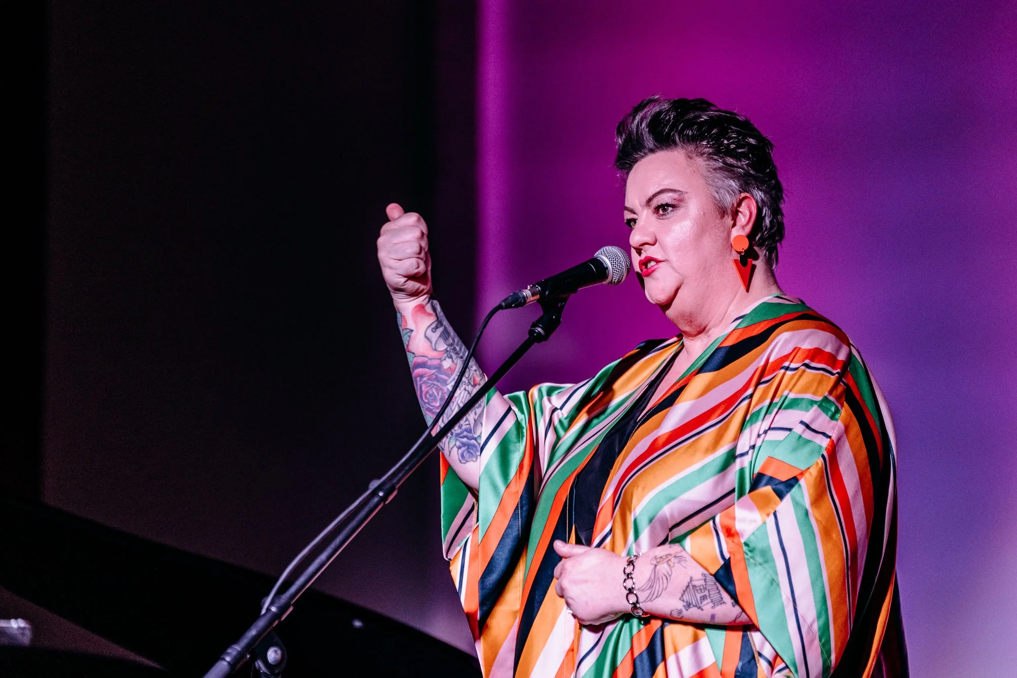Woman with short gray hair, colorful striped dress, and tattoos on her left arm, speaking into a microphone, with a purple background.