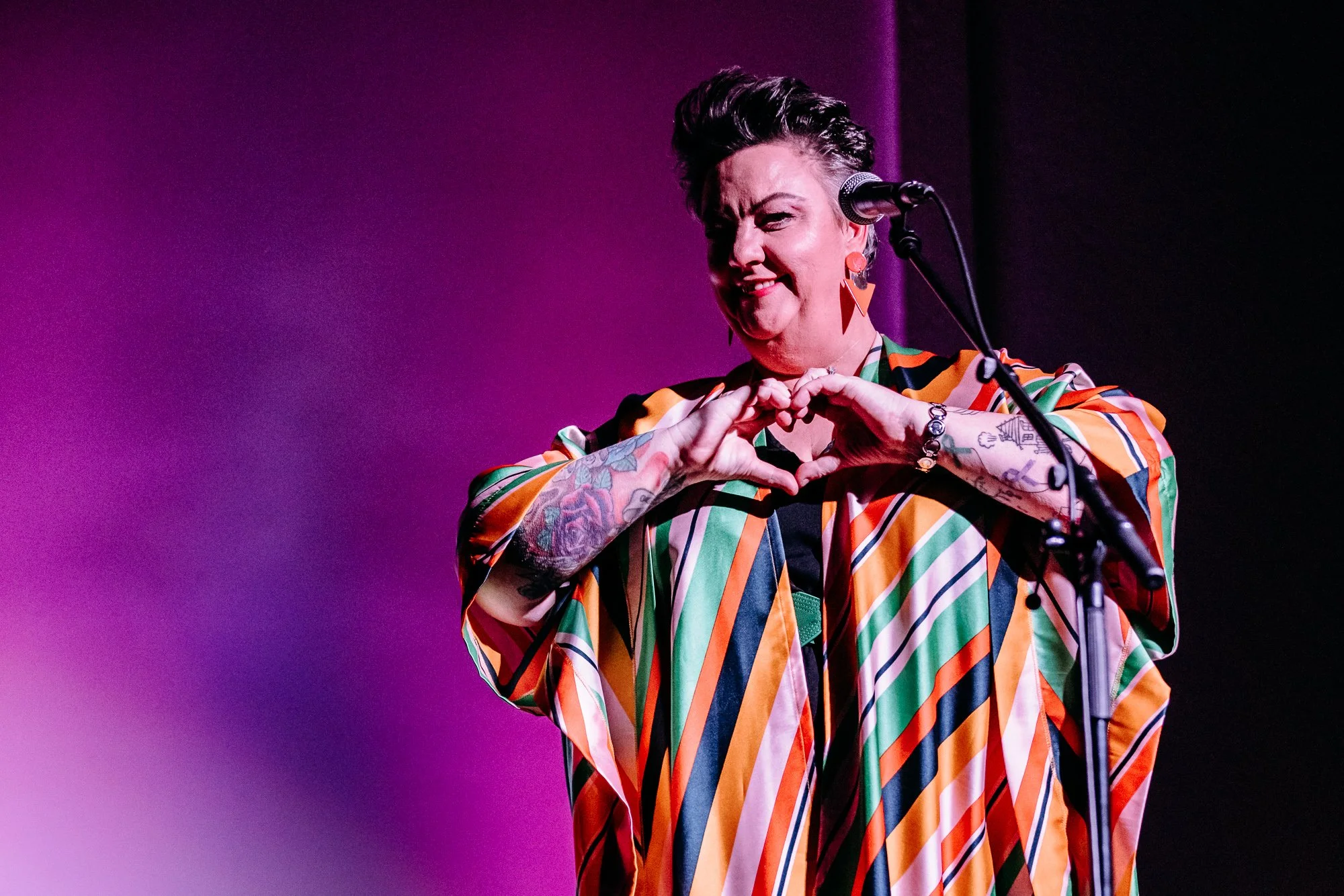 A person with short, styled hair, tattoos on their arms, and wearing large, colorful earrings making a heart shape with their hands in front of a purple background on stage with a microphone.