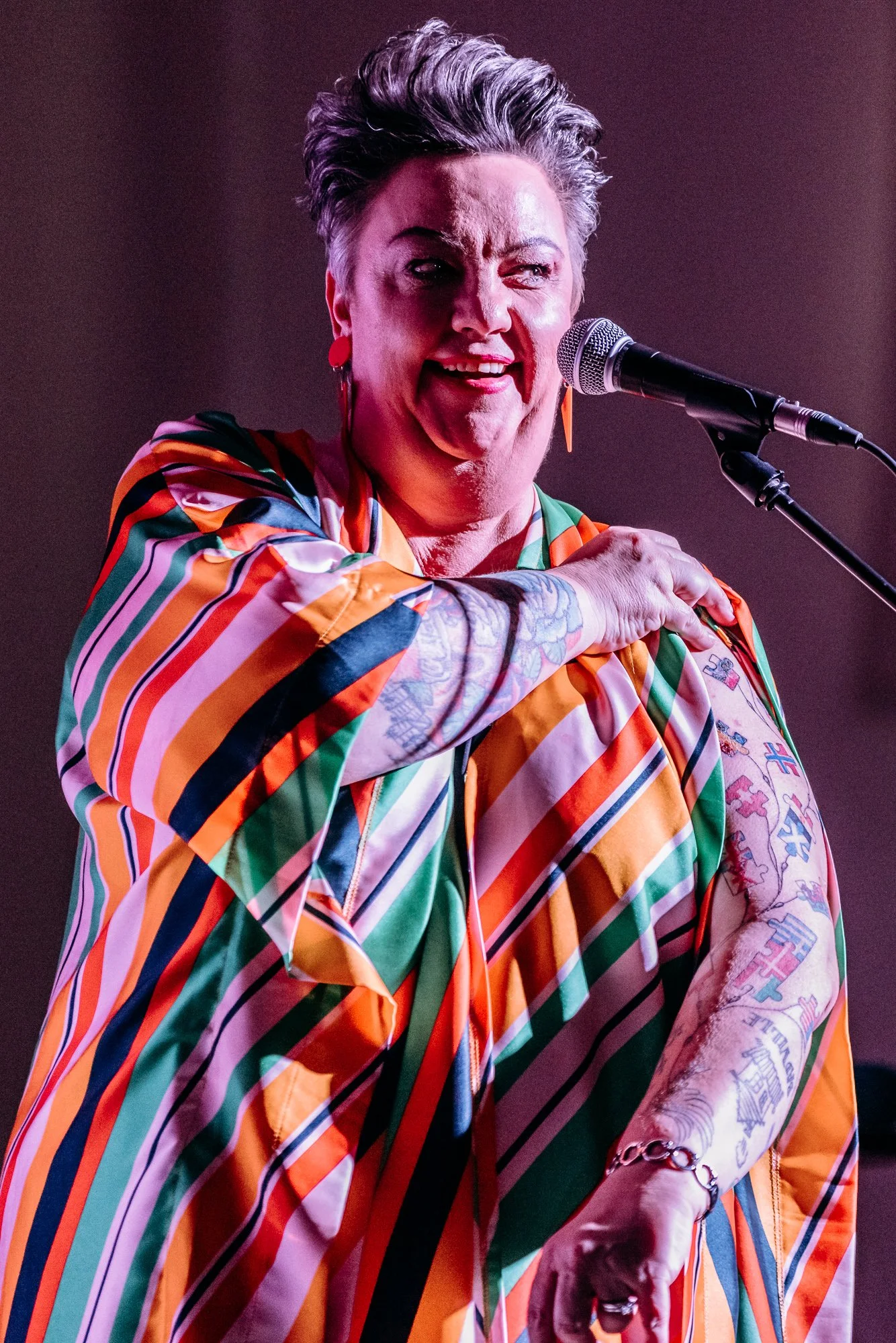 A woman with short gray hair singing into a microphone, wearing a colorful striped shirt, with tattoos visible on her arms.