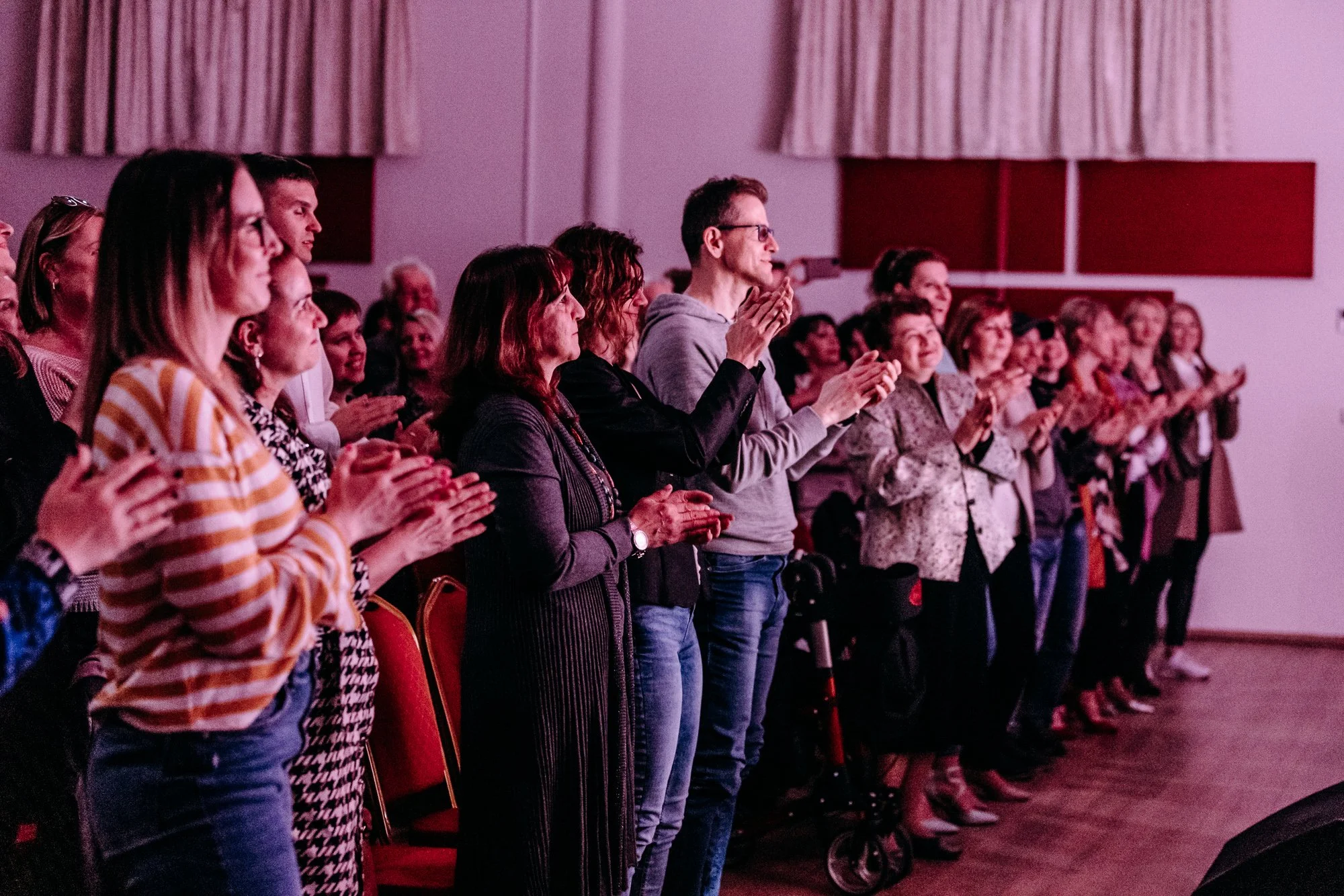 Audience standing and clapping at an indoor event with pink lighting, some wearing casual and formal attire.