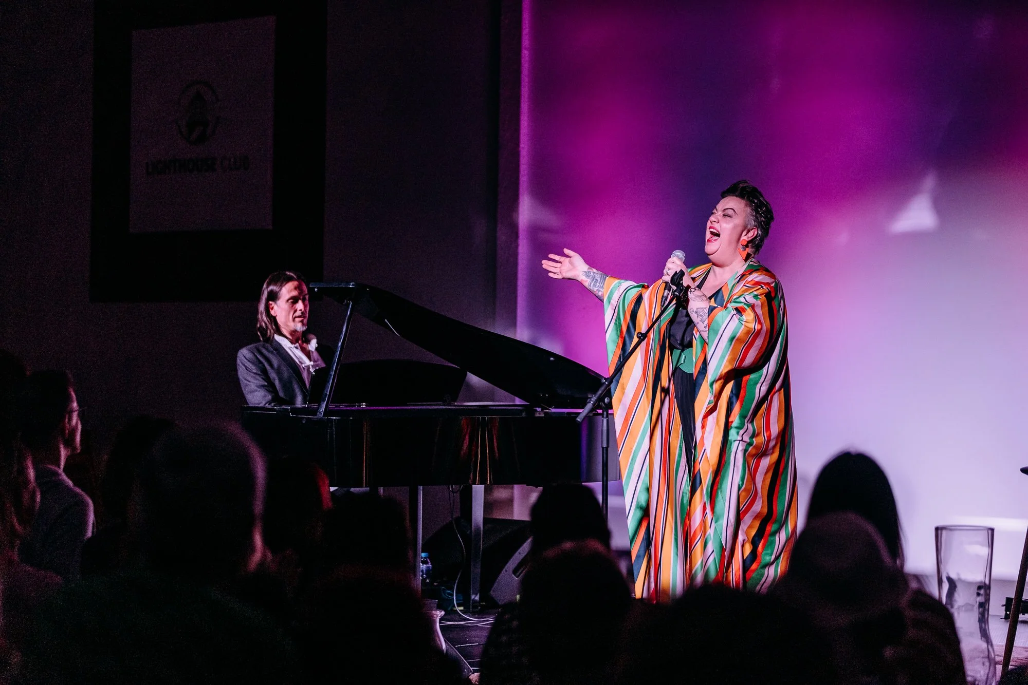 A woman singing into a microphone and gesturing with her hand on stage, wearing a colorful striped robe, with a pianist accompanying her on a grand piano in a performance venue.