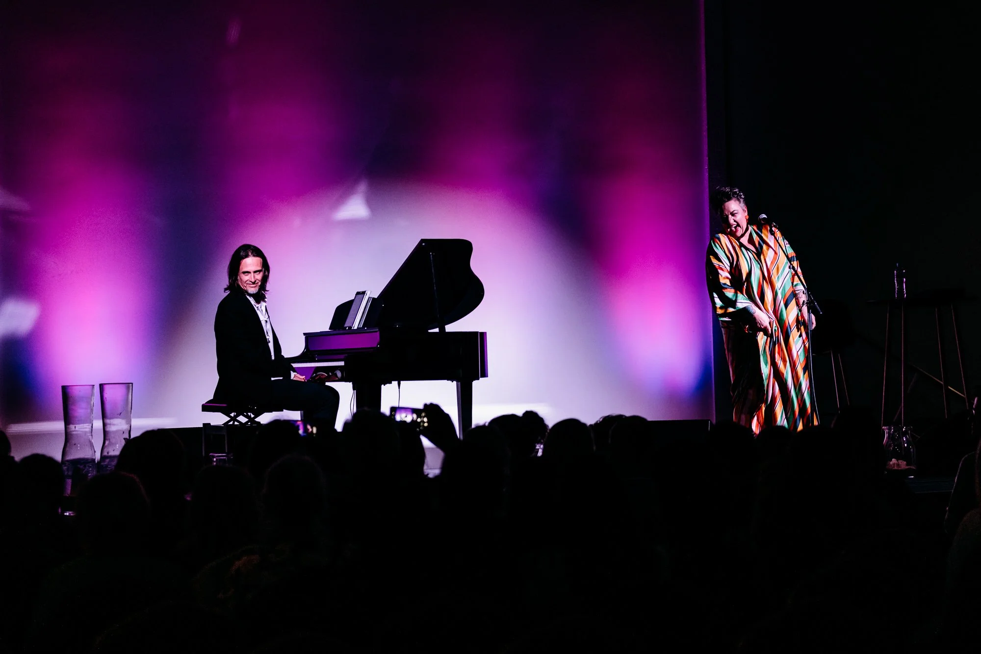 A musical performance on stage with a pianist at a black grand piano and a singer standing beside, illuminated by purple and pink stage lights, with an audience watching.