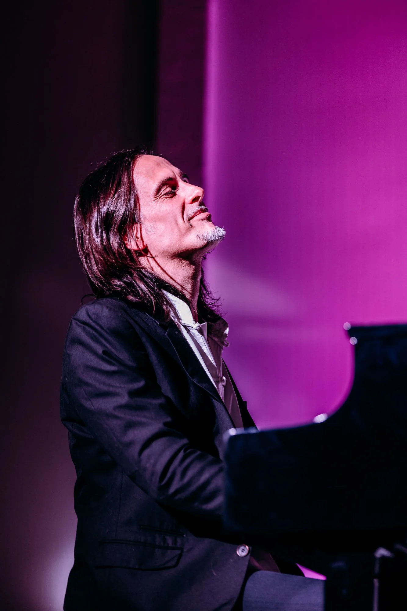 A man with long hair and a goatee playing a piano, wearing a black jacket, with purple stage lighting in the background.