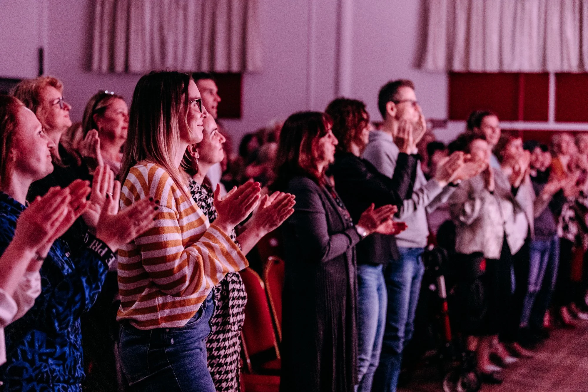 An audience standing and clapping at an indoor event, some wearing glasses and casual clothing, with pink lighting.