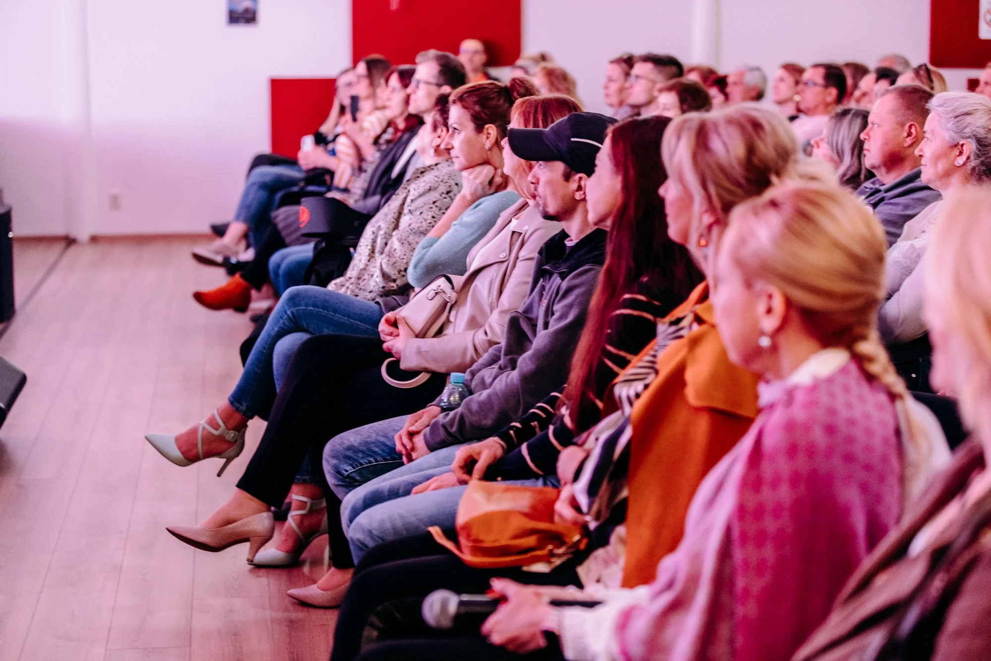 Audience members sitting in a conference room, attentively listening to a speaker, with some taking notes and others holding water bottles.