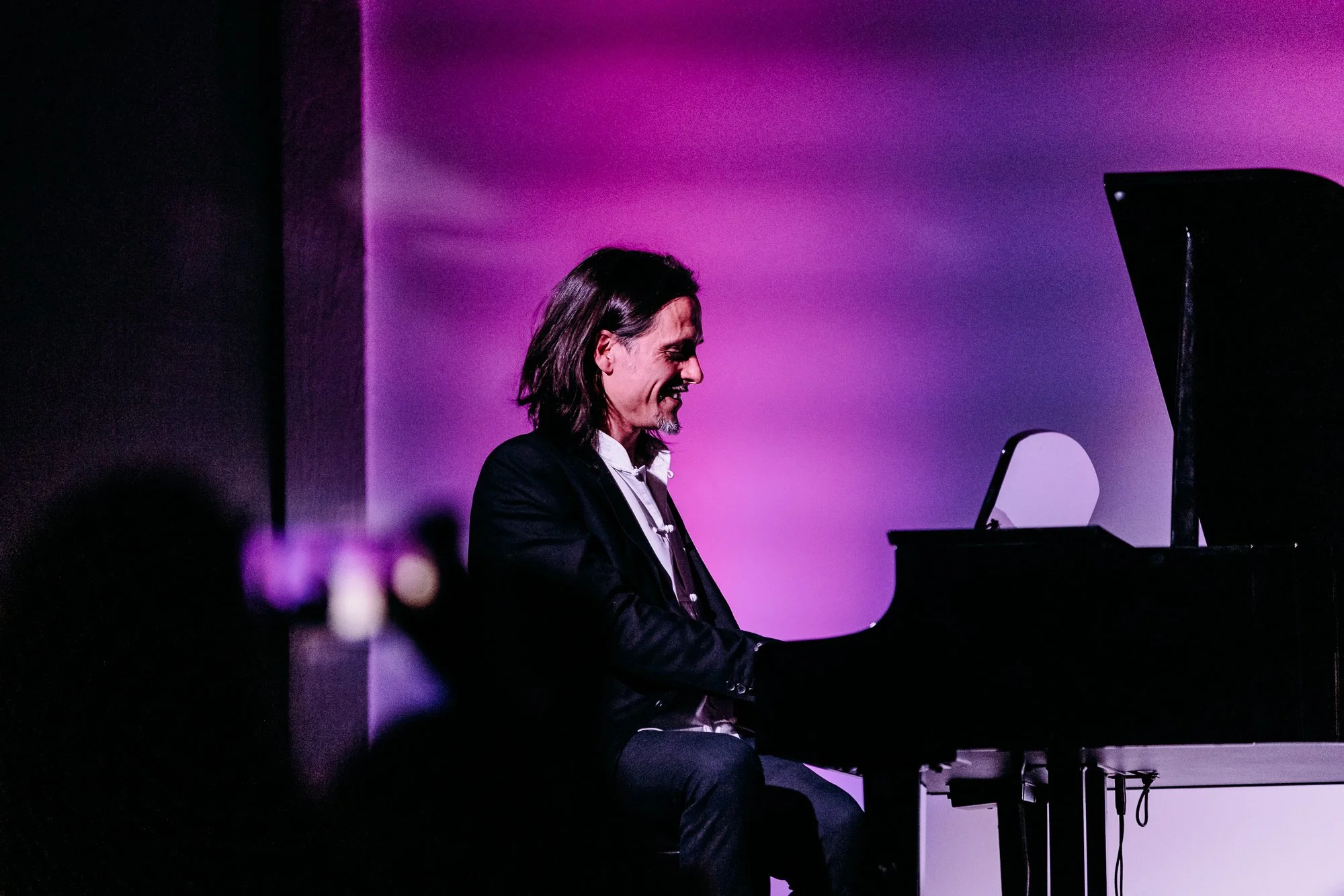 A man playing a grand piano on stage with purple lighting, smiling and dressed in a dark blazer.