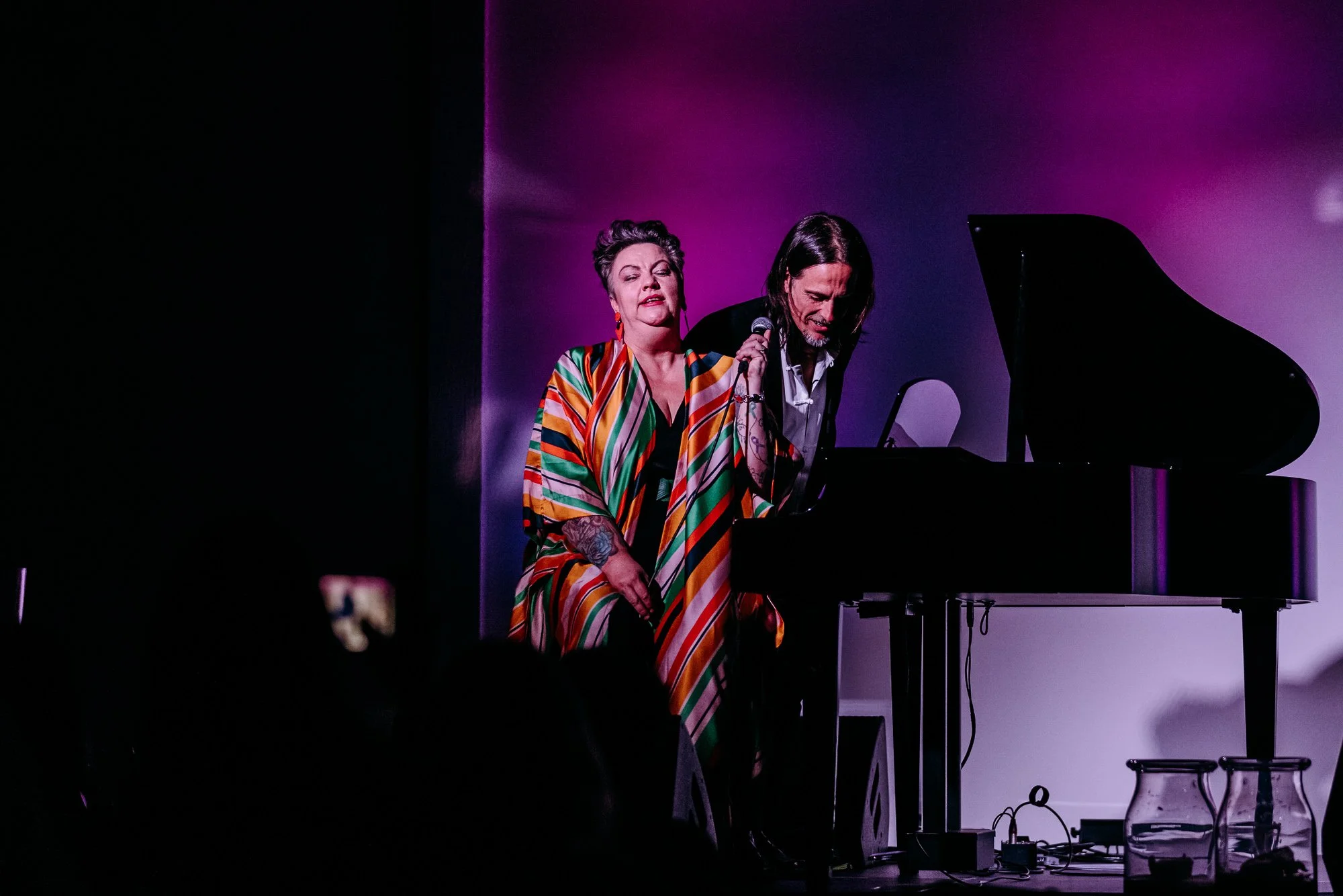 A woman singing into a microphone beside a man at a black grand piano on a stage with purple lighting.