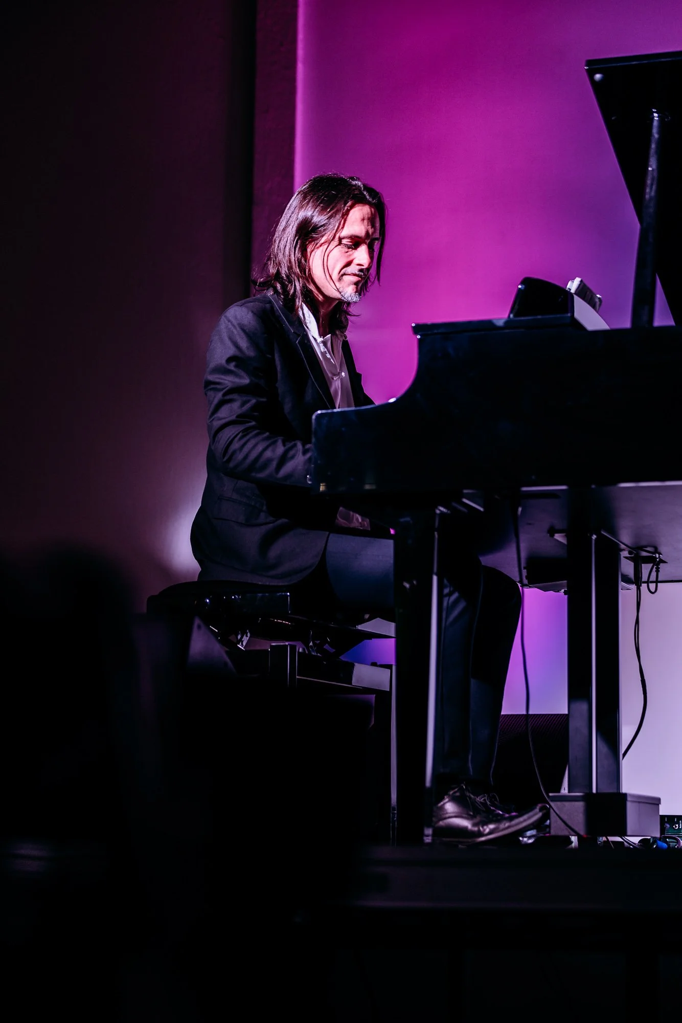 Man playing grand piano on stage under purple lighting.
