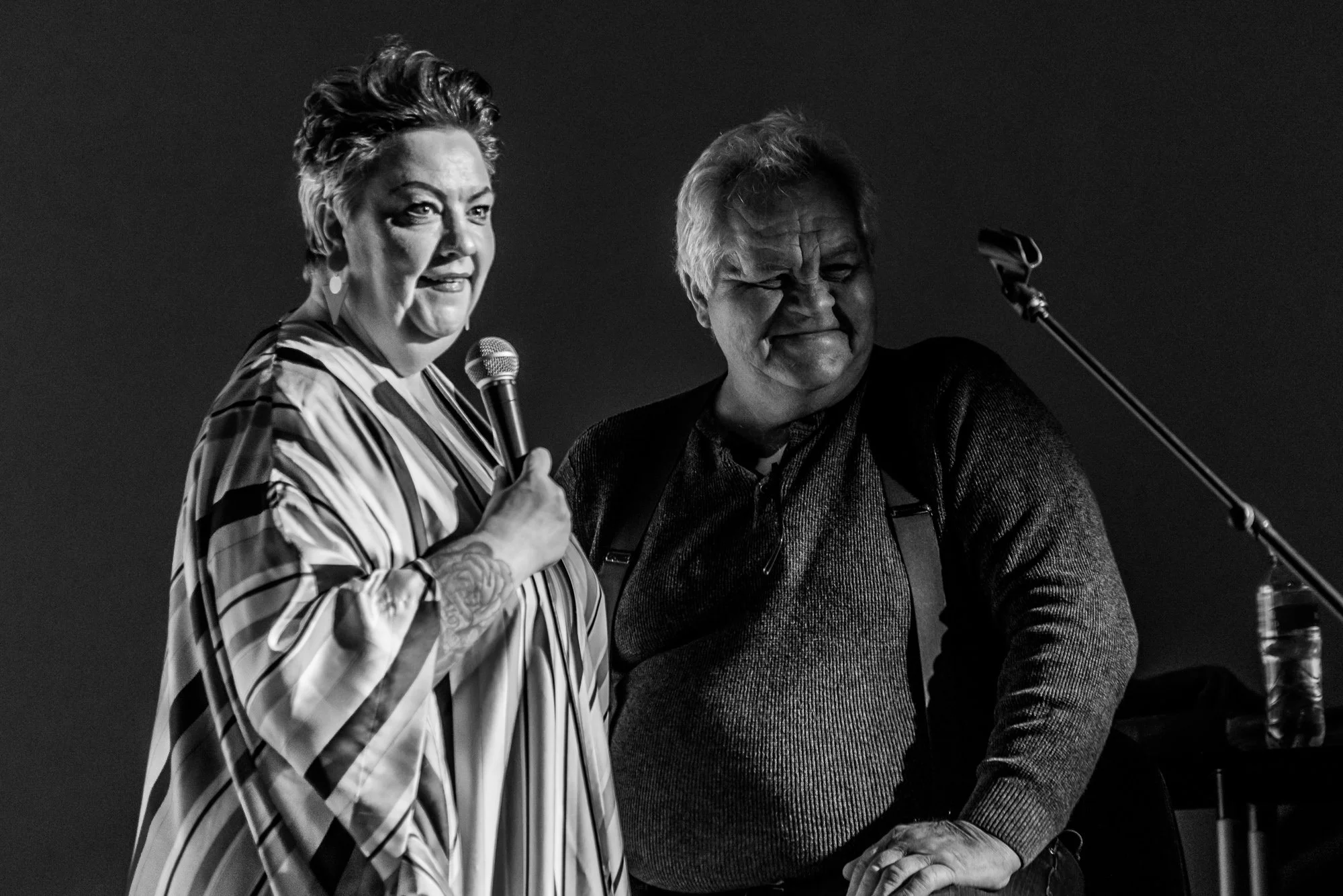 A woman with short, styled hair holding a microphone and speaking, standing next to a man with glasses and gray hair, who is smiling and has his hand on his hip, in a black and white photo.