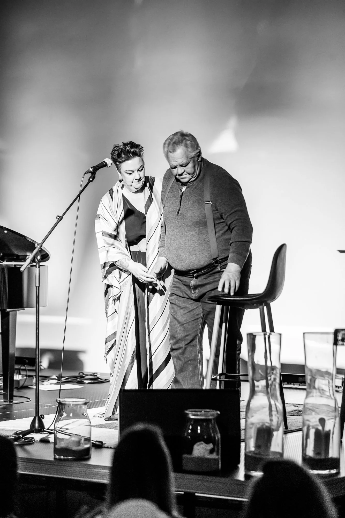 Black and white photo of a woman and a man on stage. The woman is wearing a striped dress, and the man is dressed casually with suspenders. They are standing close together, looking down, with a microphone on a stand nearby.