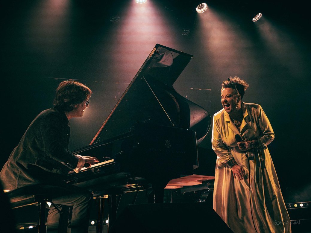 A pianist playing a grand piano with a singer performing passionately next to him, both on stage under warm lighting.