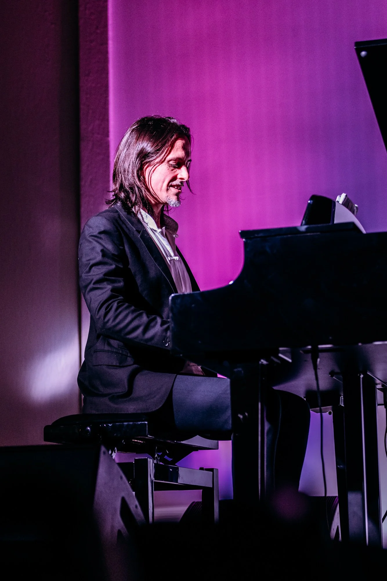 A man with shoulder-length dark hair, wearing a black suit jacket and a patterned shirt, playing a grand piano on stage with purple lighting.