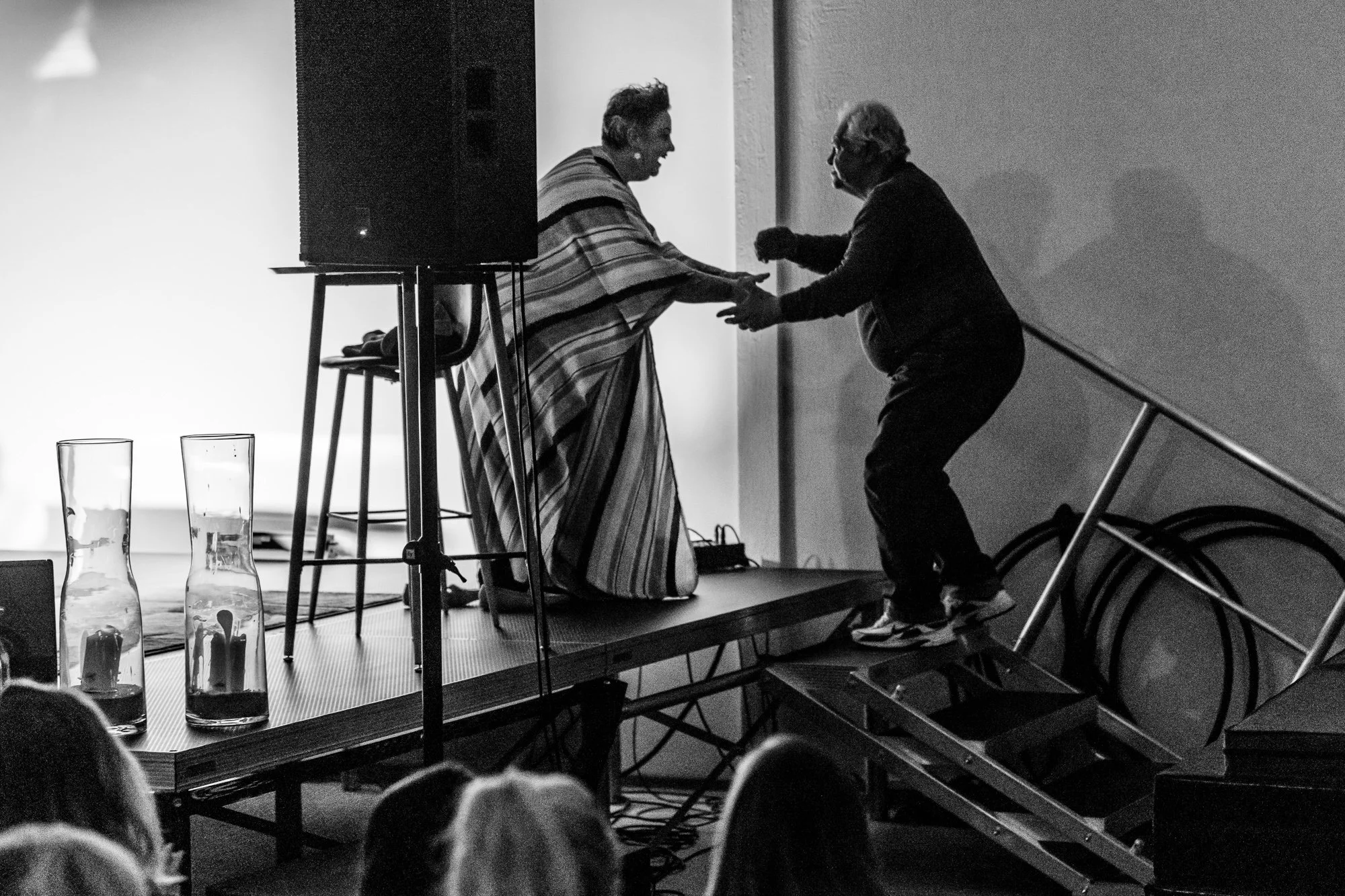 A person draped in a striped blanket dancing with an older man on a small elevated stage, while an audience watches.