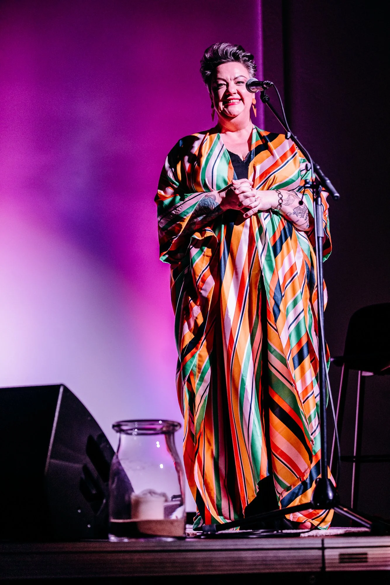 A woman with short hair and tattoos on her arms, wearing a colorful, striped dress, is standing on a stage in front of a microphone, smiling and making a playful face. There is a glass jar with a candle and some books on the floor in front of her, wi