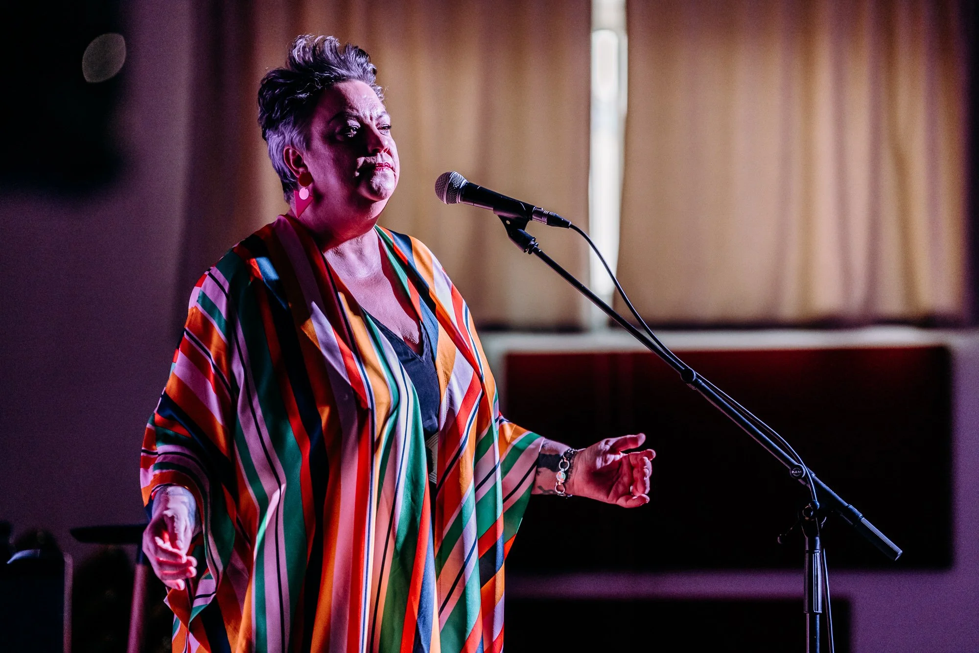 A woman with short gray hair singing into a microphone on stage, wearing a colorful, striped, loose-fitting top, in a room with wood panel walls and a window with curtains.