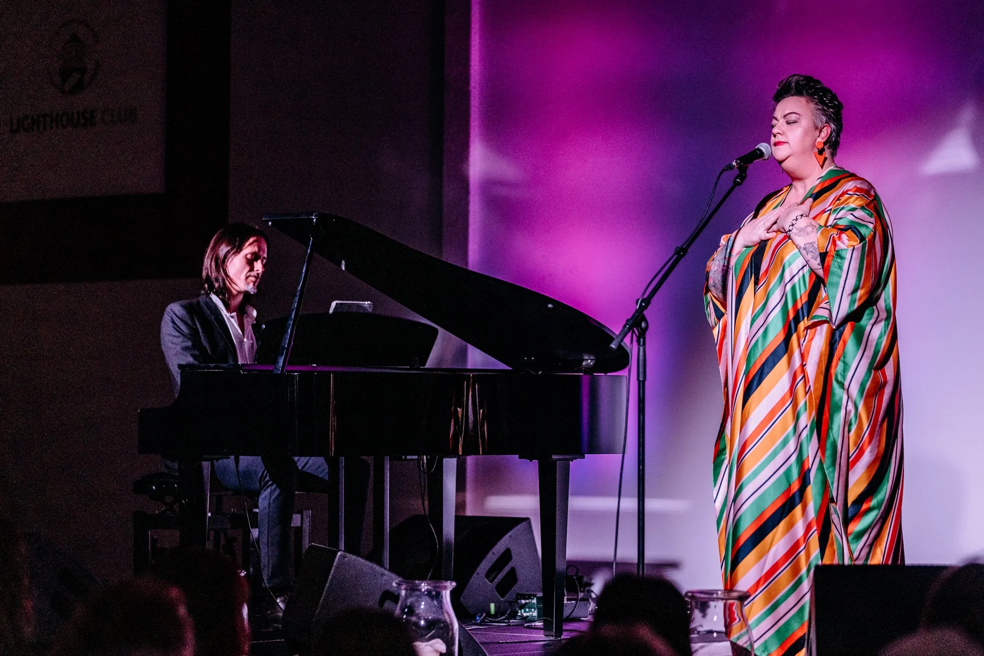 A woman in a colorful striped dress sings into a microphone while a pianist plays on a black grand piano behind her. They are on a stage with purple lighting and a sign that says 'Lighthouse' in the background.