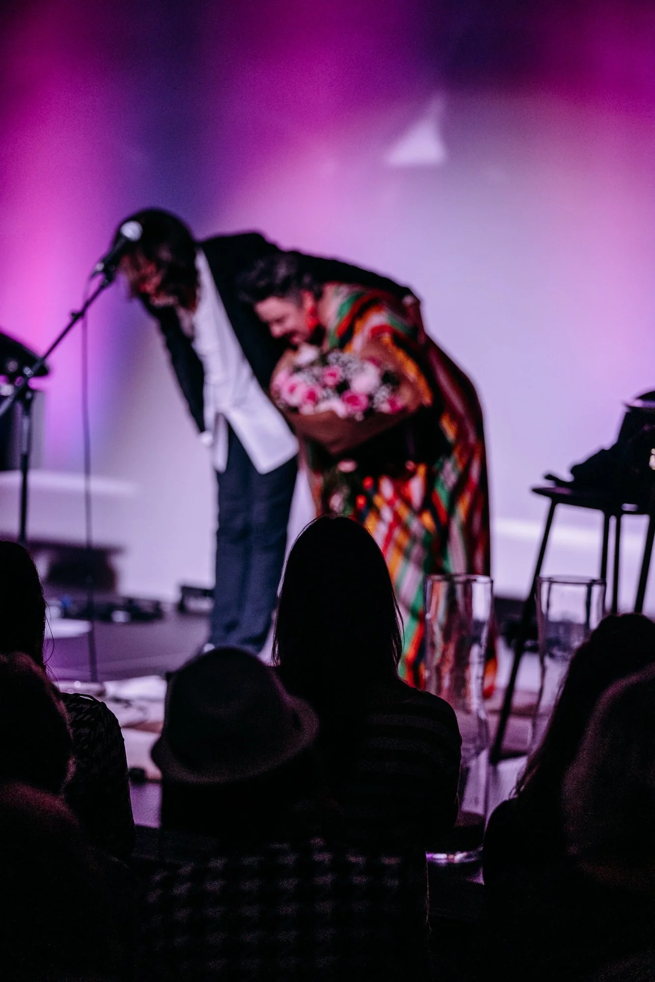 Two performers bowing on stage after a performance, one holding a bouquet of flowers, in front of an audience.