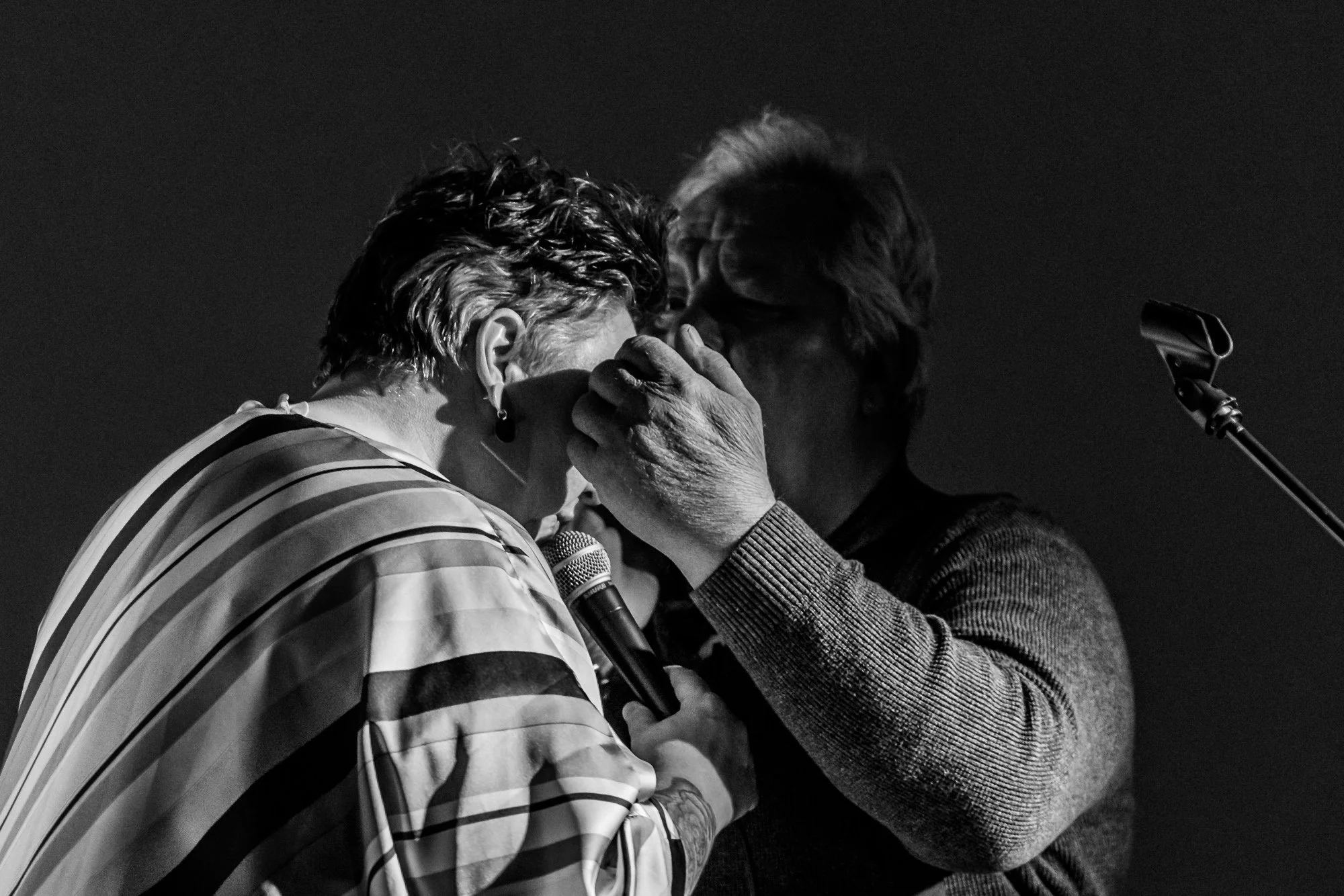 A man and woman share an intimate moment on stage, with the man gently holding the woman's face as they kiss. The scene is in black and white, with a microphone in the woman's hand and a microphone stand nearby.