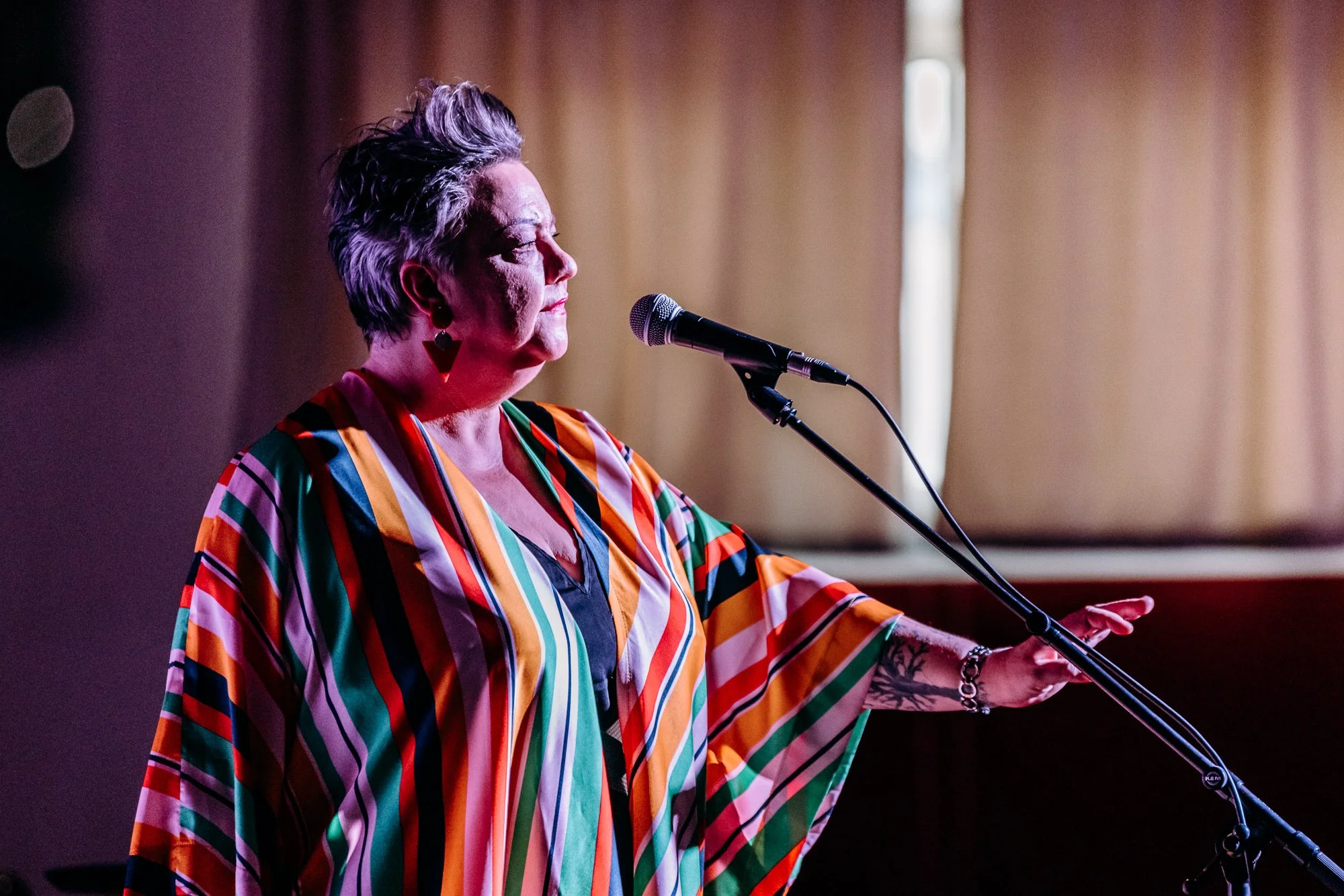 An older woman with short gray hair, wearing a colorful striped dress and large earrings, standing at a microphone with her hand outstretched, in front of beige curtains.