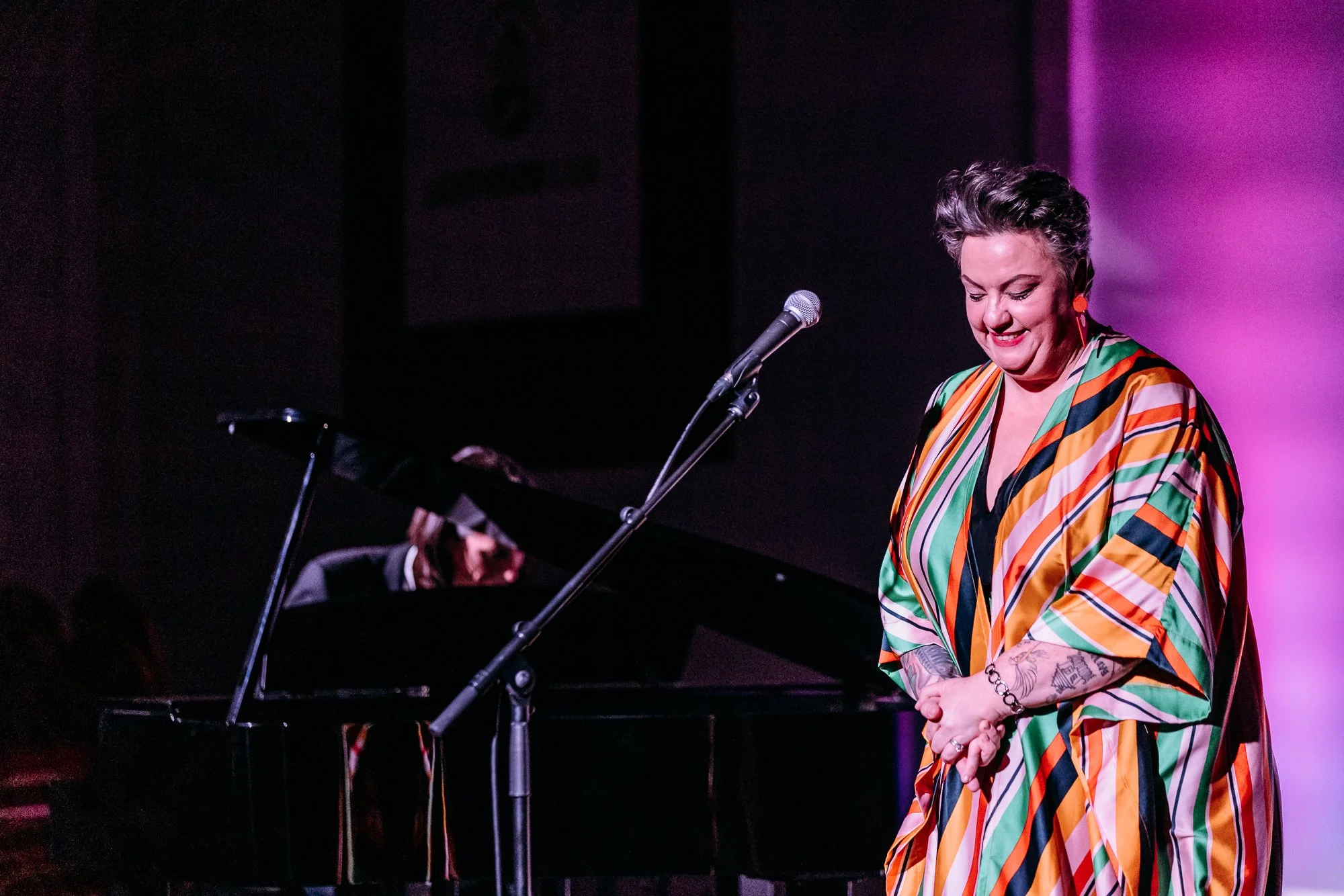 A woman in a colorful, striped dress smiles on stage while bowing, with a grand piano in the background and a male pianist partially visible playing the piano.