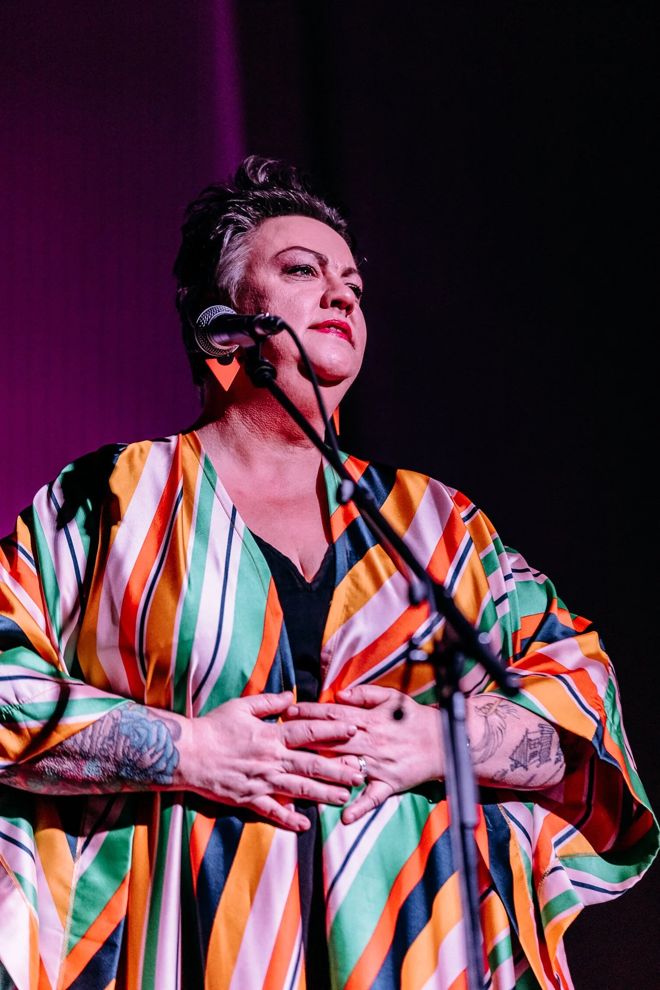 A woman with short dark hair and tattoos on her arms, wearing a colorful striped jacket, standing behind a microphone with her hands on her chest, on a stage with a purple background.
