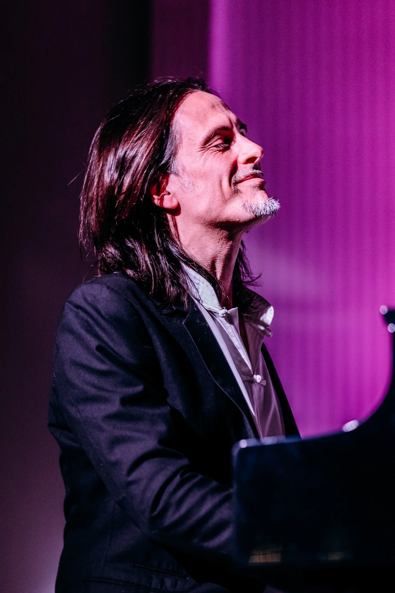 A man with long dark hair and a goatee playing a piano, with a purple background.
