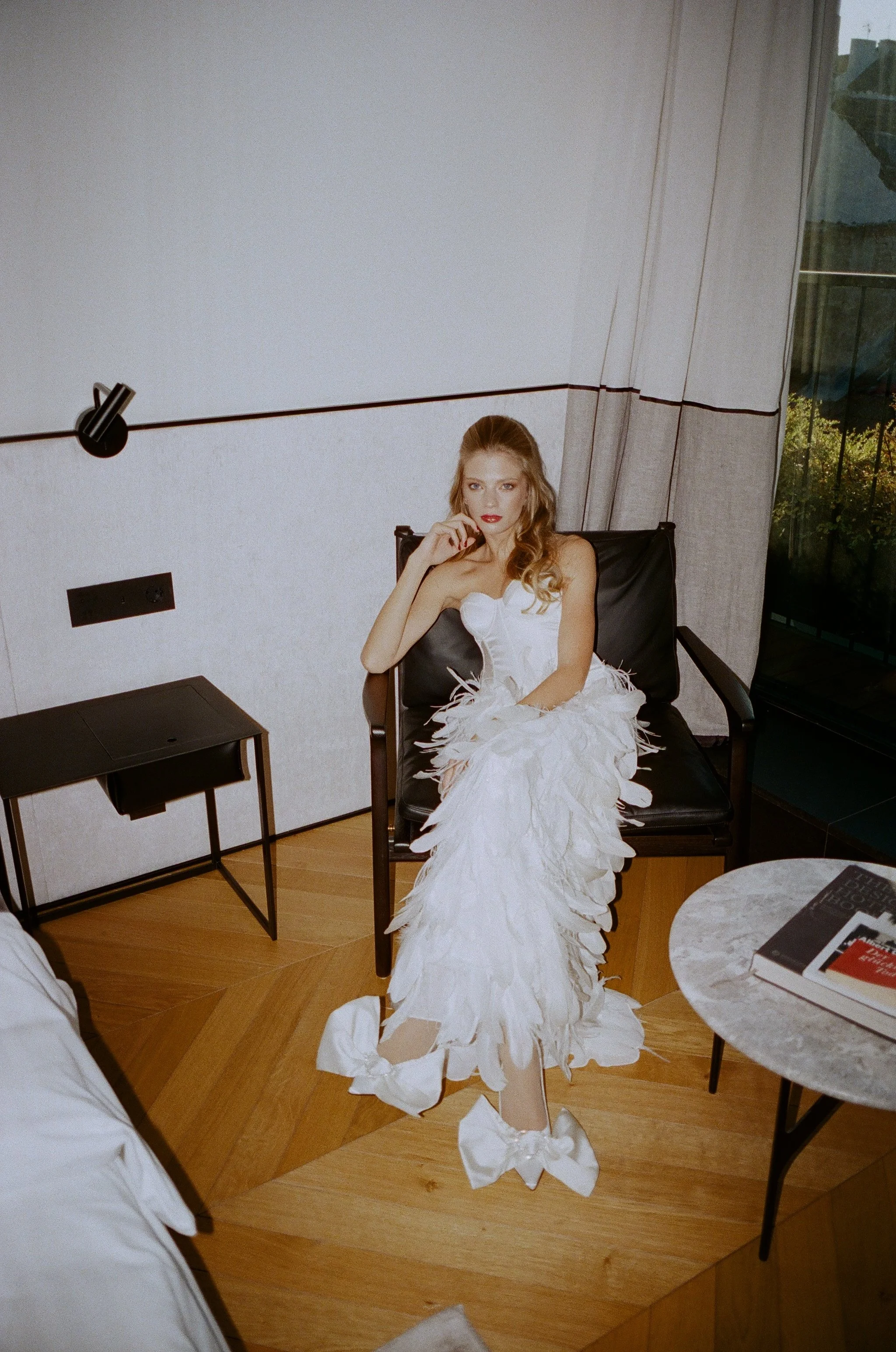 bride in a strapless feathered wedding gown with satin bow heels, seated in a minimalist interior · editorial bridal style · film photography by radostina boseva