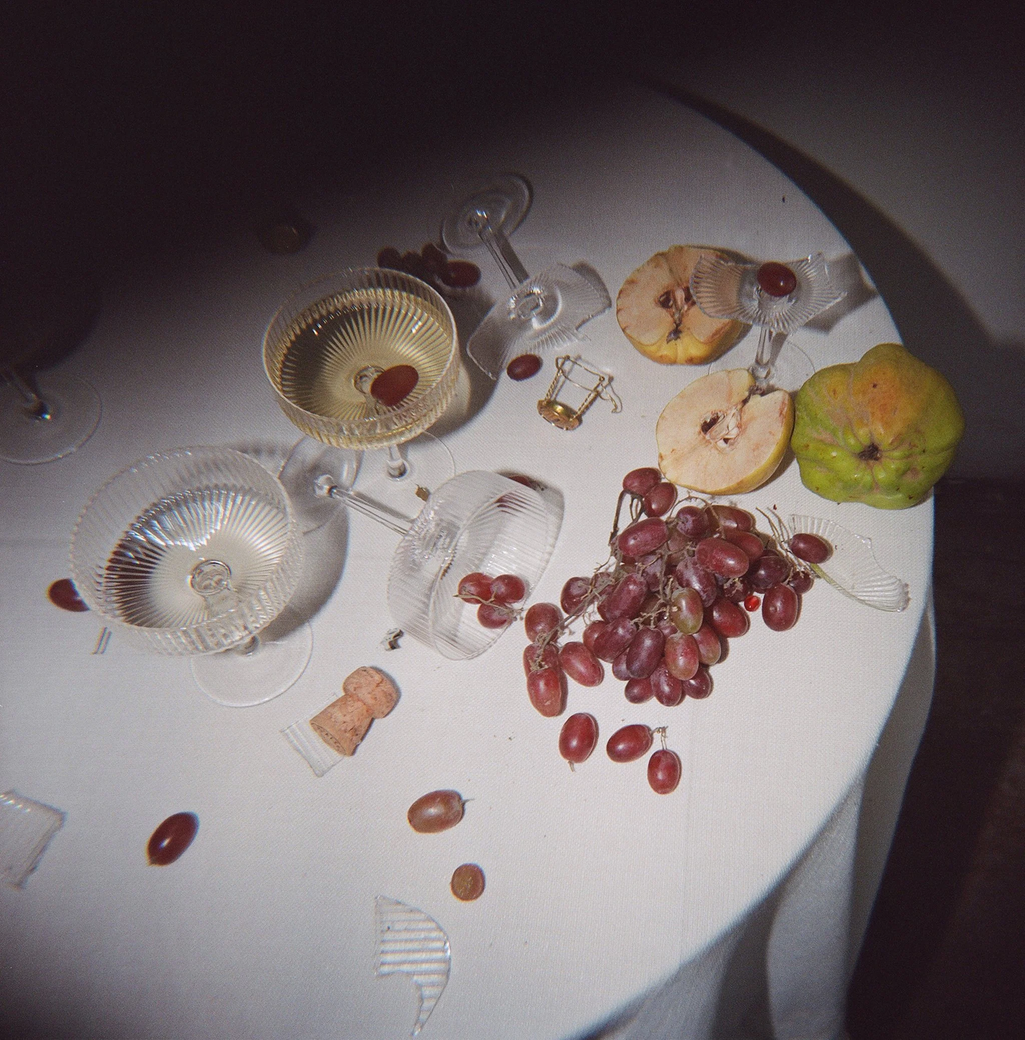 overhead flash photograph of a table after the toast — coupe glasses, scattered grapes, quince, and a champagne cork, on film