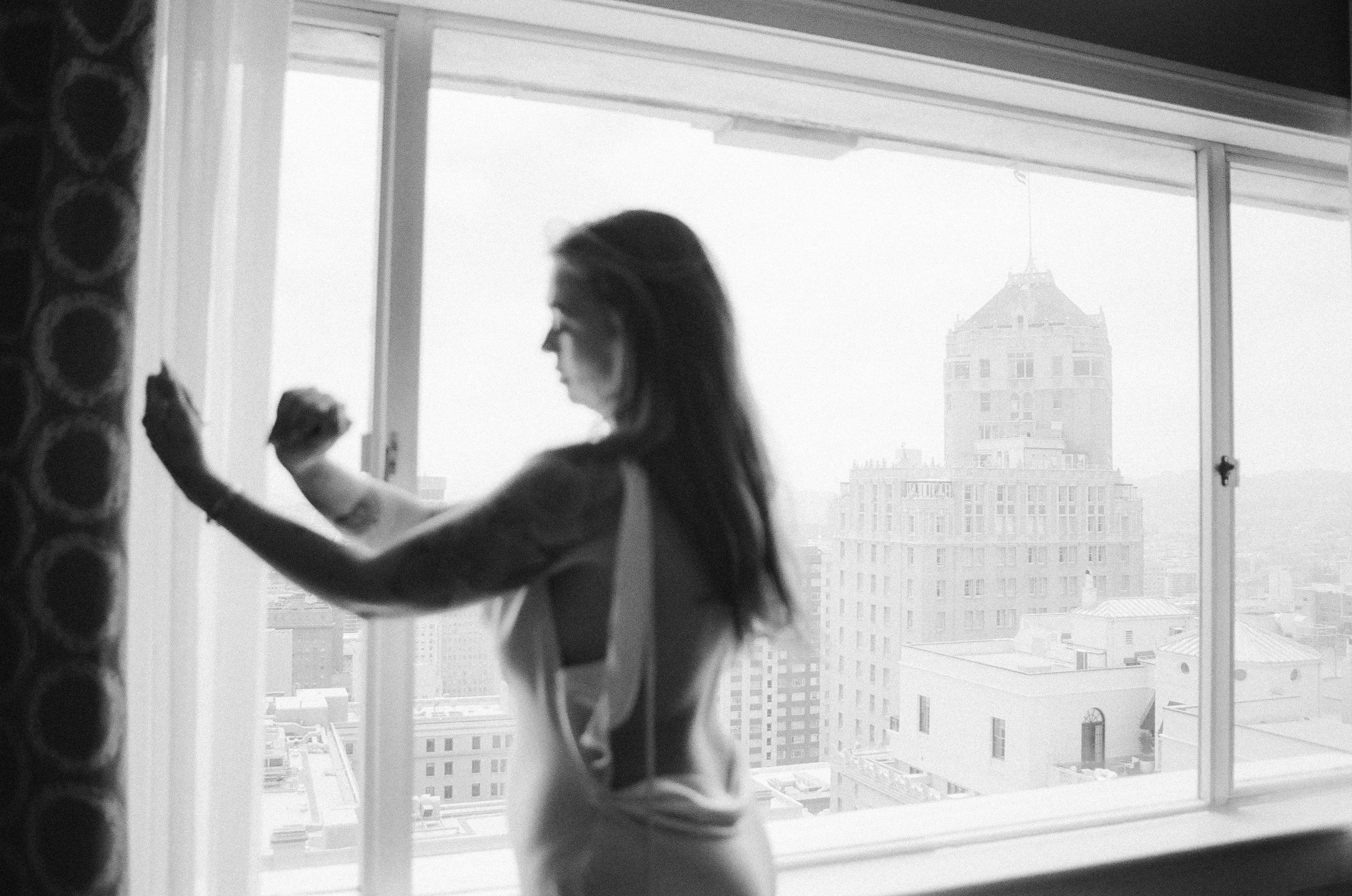 bride reaching toward the window as a city tower emerges through the fog · fairmont hotel san francisco getting ready · documentary film photography by radostina boseva