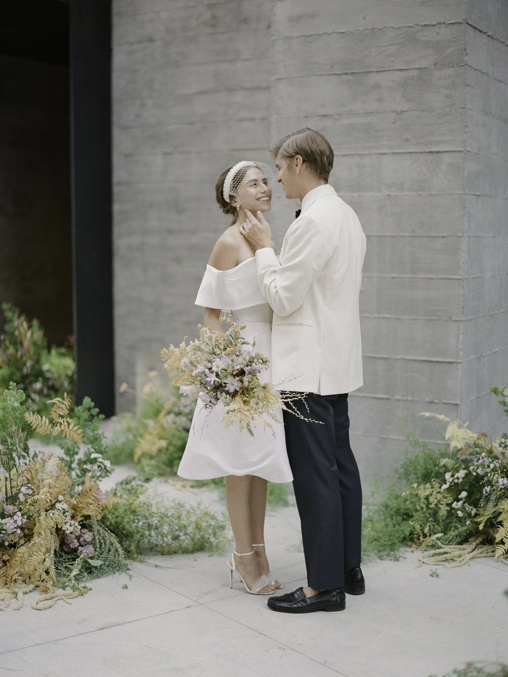 couple in a short off-shoulder dress and ivory jacket embracing against raw concrete architecture, wildflowers underfoot · modern minimalist wedding style · editorial film photography by radostina boseva