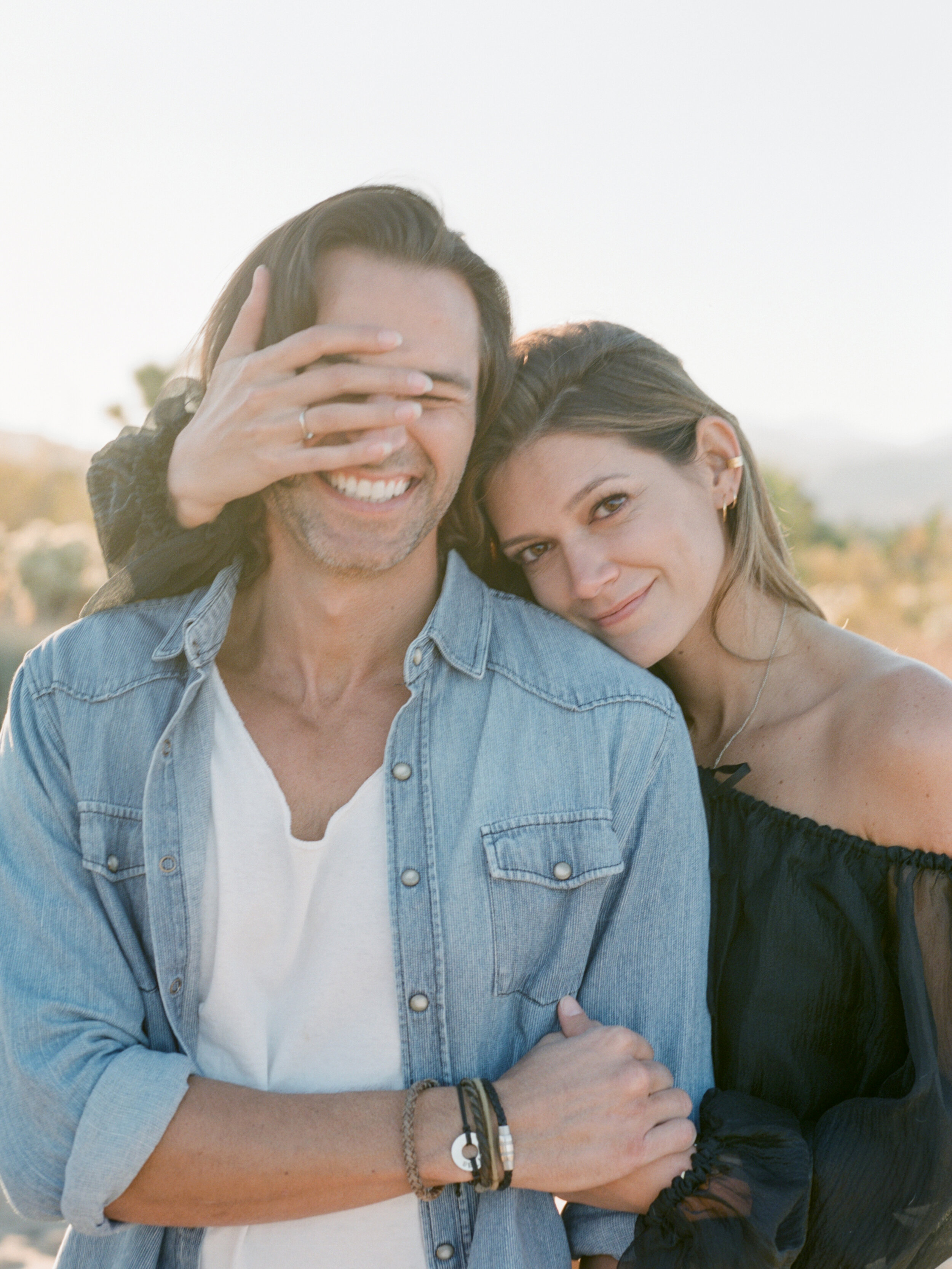 a couple at golden hour in the high desert during a joshua tree engagement session — she rests against his shoulder while his hand covers his eyes mid-laugh, her ring in frame, photographed on film by radostina boseva