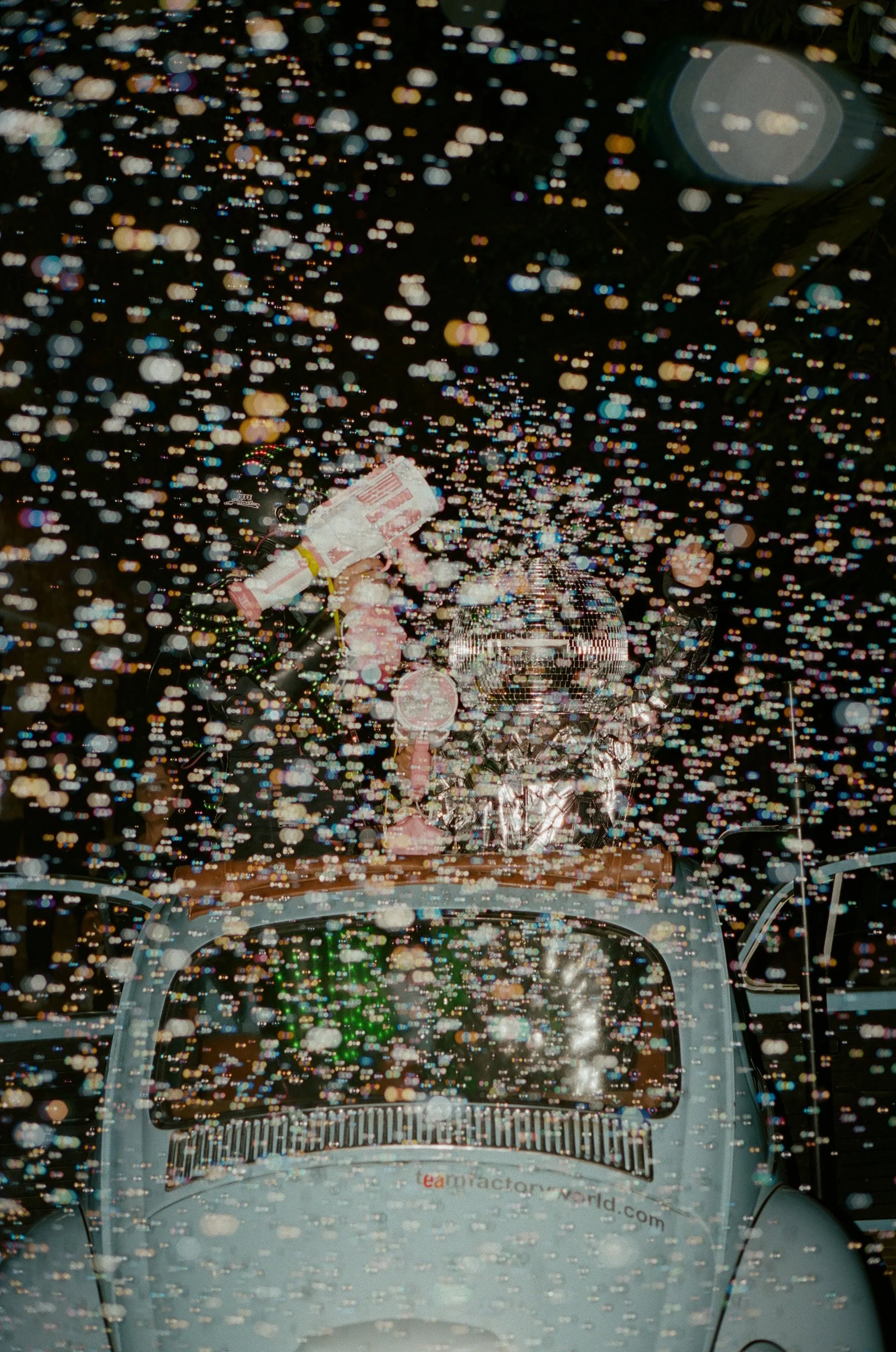 newlyweds leaving in a vintage volkswagen beetle through a storm of bubbles, one wearing a disco ball helmet, at night on film