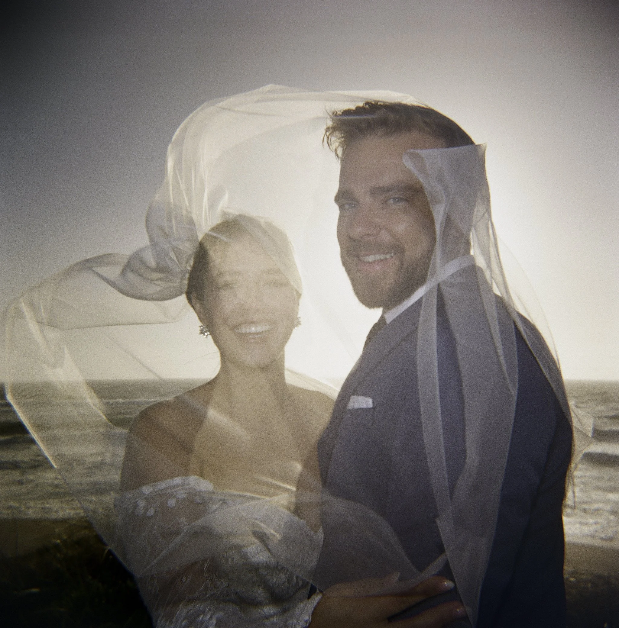 couple embracing as the veil catches the wind at golden hour, ocean behind them · big sur california elopement · documentary film photography by radostina boseva