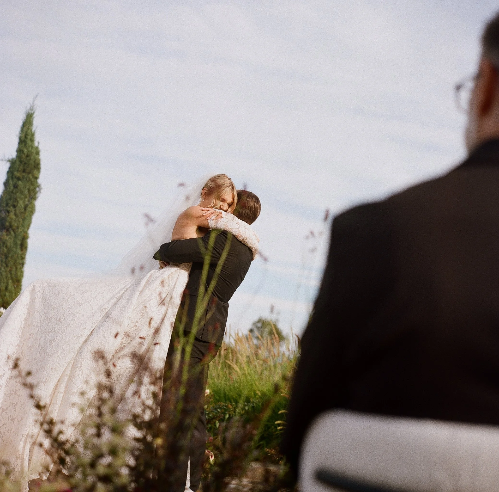 groom lifting the bride in an embrace during the ceremony, seen over a guest's shoulder, on film