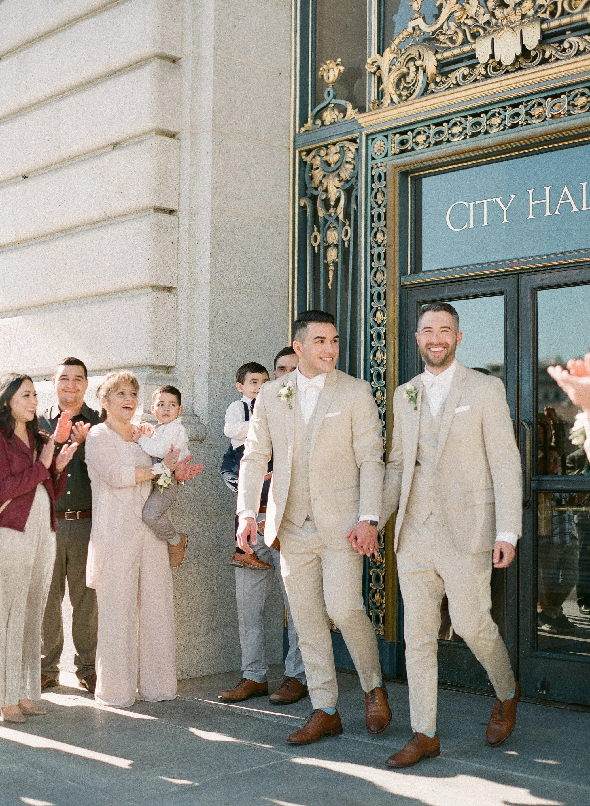 Two grooms in matching champagne suits exiting San Francisco City Hall to cheering guests, film wedding photography by Radostina Boseva