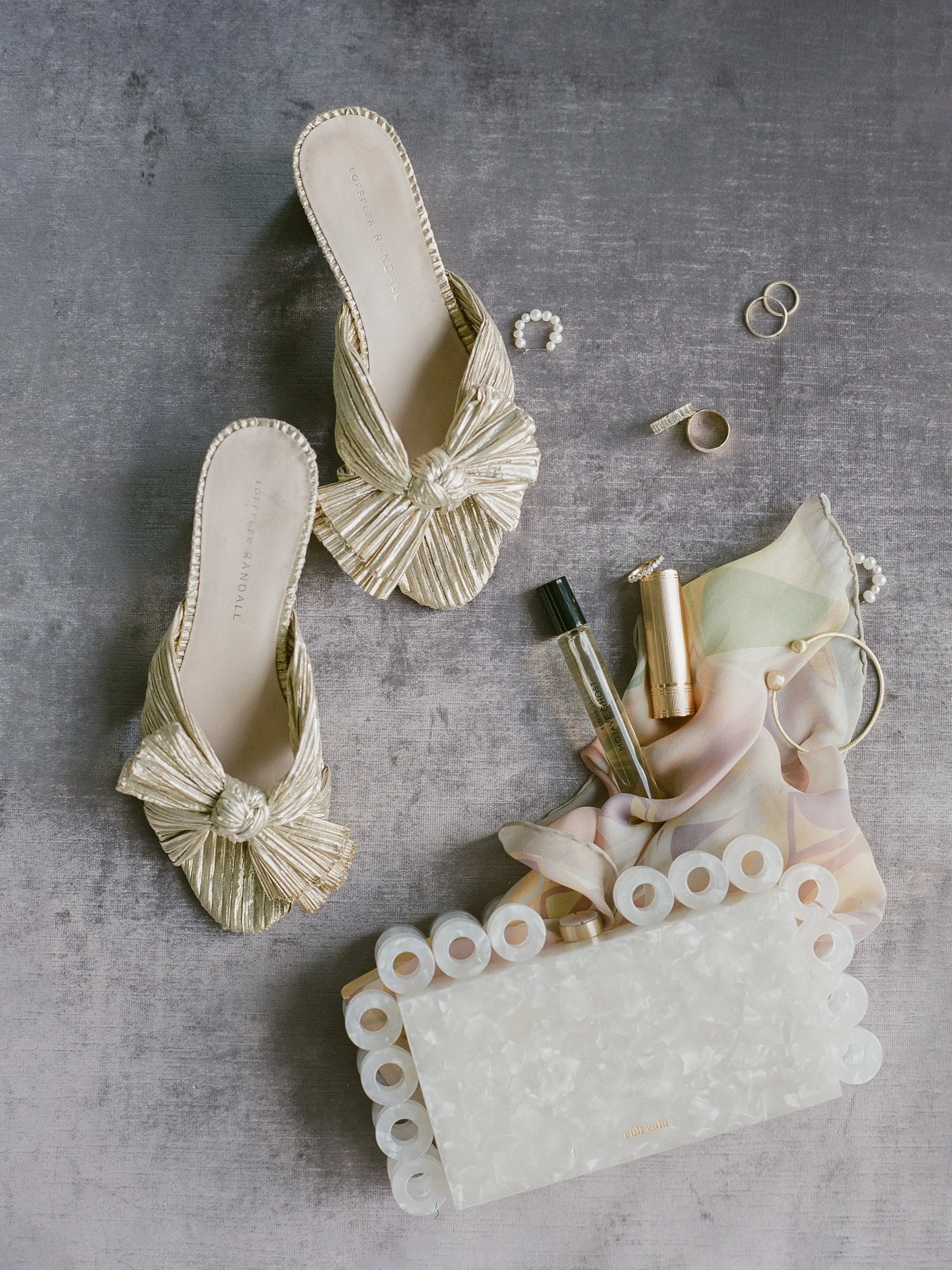 Bridal detail flat lay — gold knot heels, wedding bands, pearl ring, acrylic clutch and silk scarf, film photography by Radostina Boseva