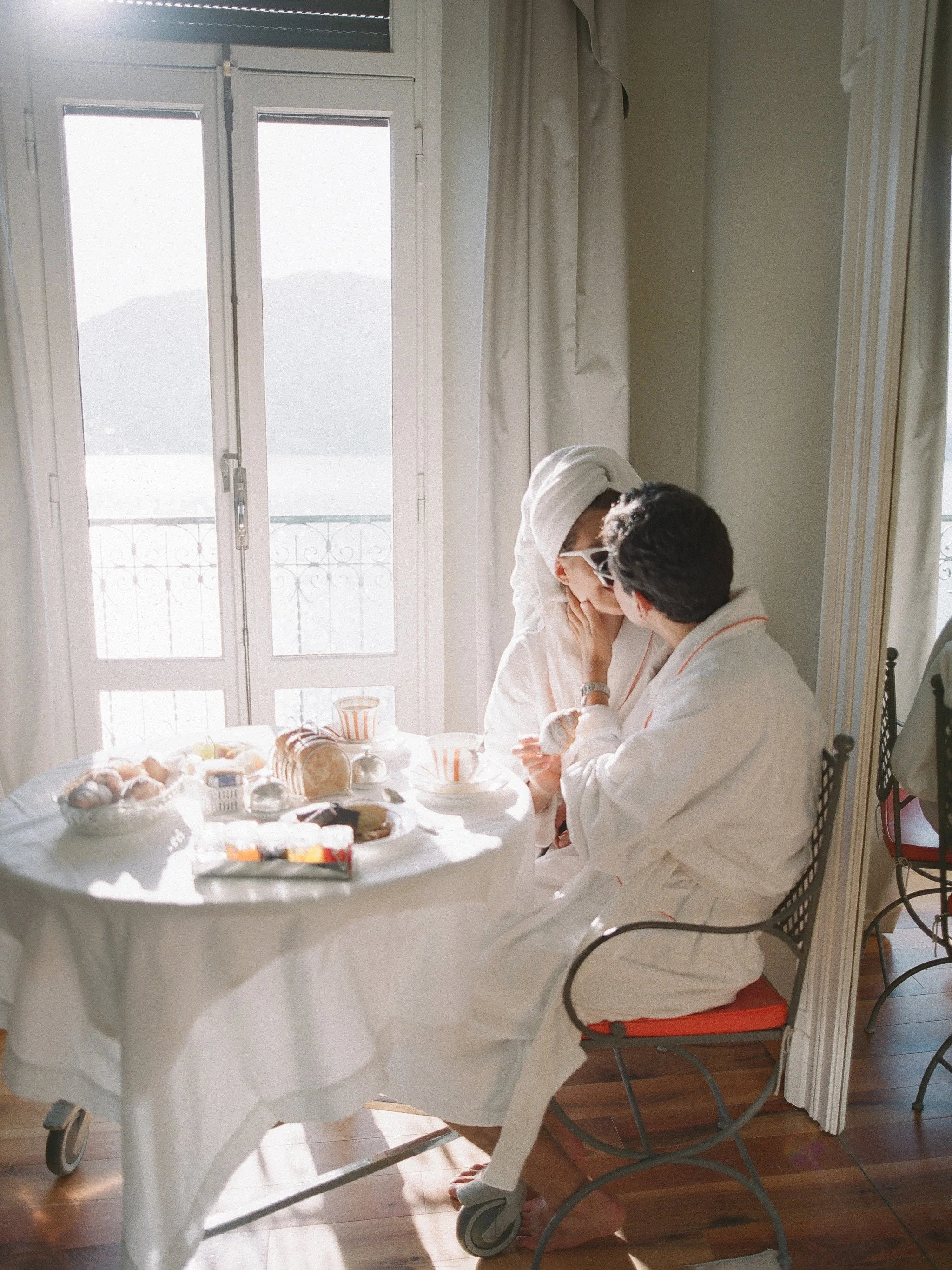 couple sharing breakfast in white robes by a window overlooking lake como · italy honeymoon morning · documentary film photography by radostina boseva