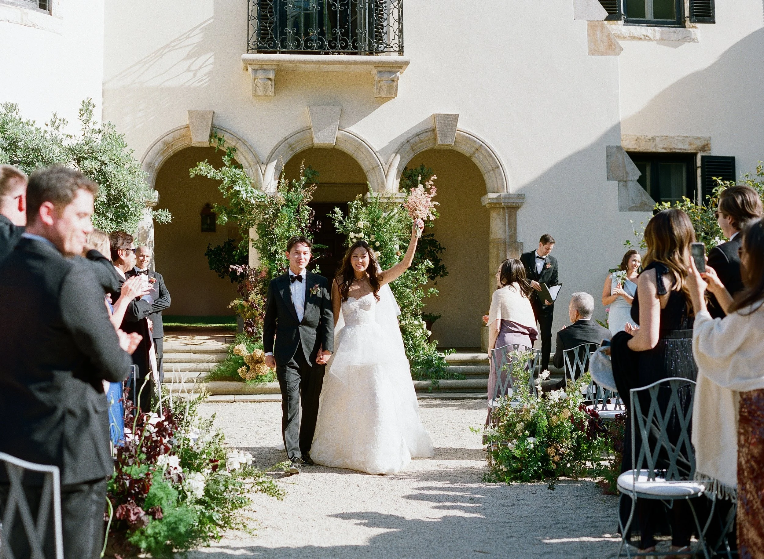 Bride and groom recessional through the courtyard at Stonepine Estate Chateau Noel, bride raising bouquet through floral aisle lining, film wedding photography by Radostina Boseva Carmel Valley