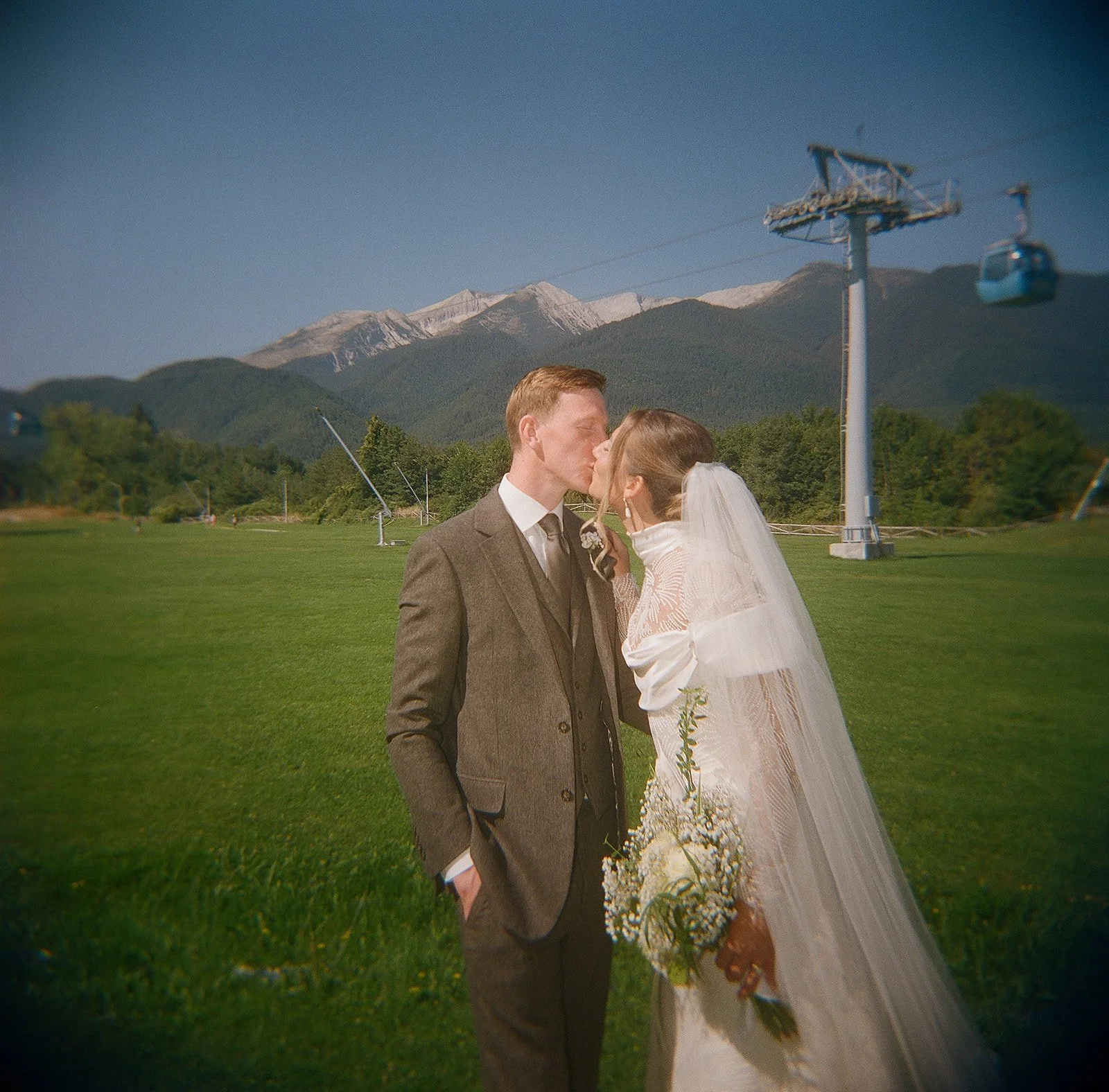 Bride and groom kissing in alpine meadow with snow-capped mountains and cable car, destination wedding photographer Austria
