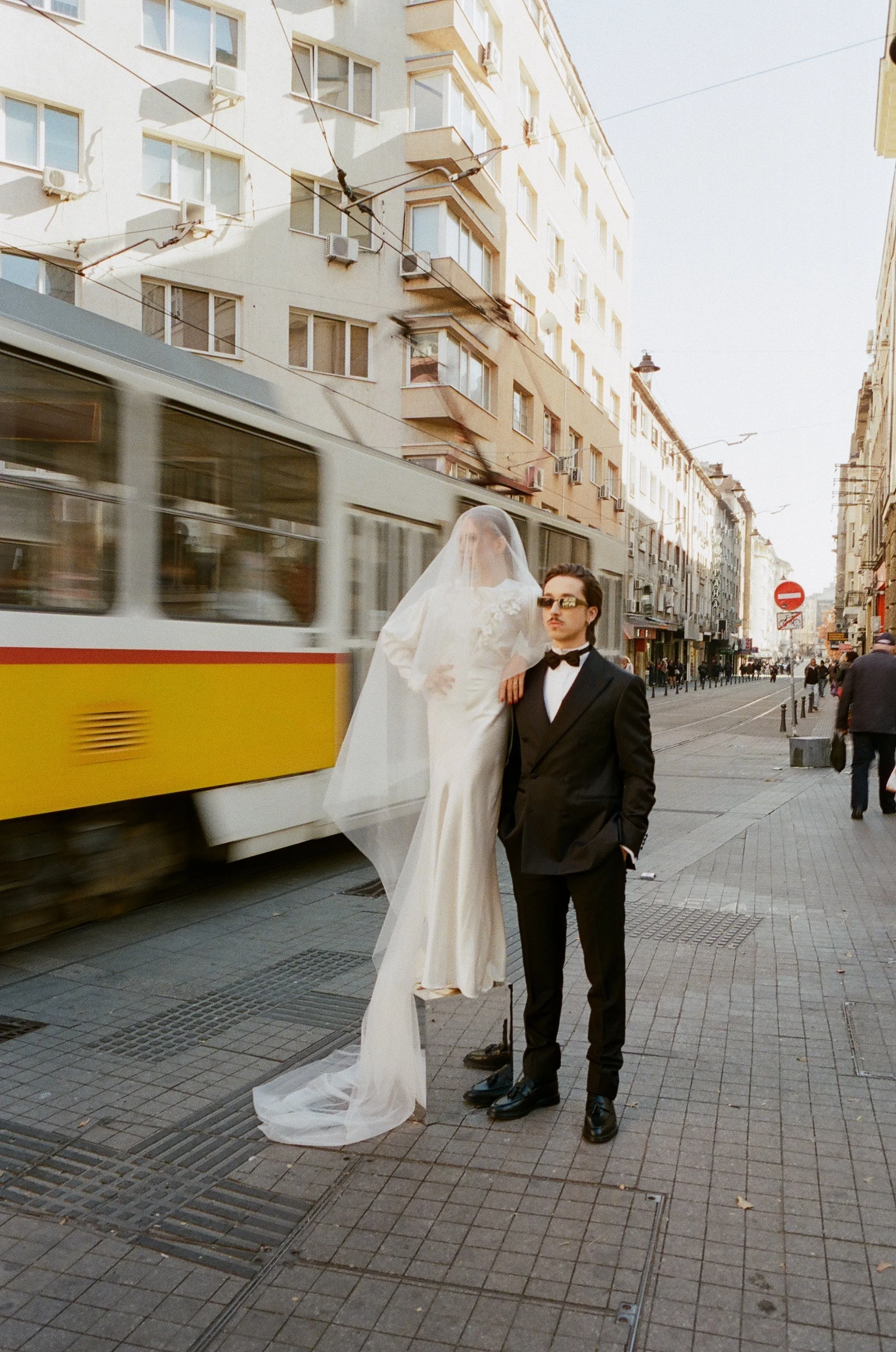 bride and groom pausing on a european city street as a tram passes · destination elopement · documentary film photography by radostina boseva