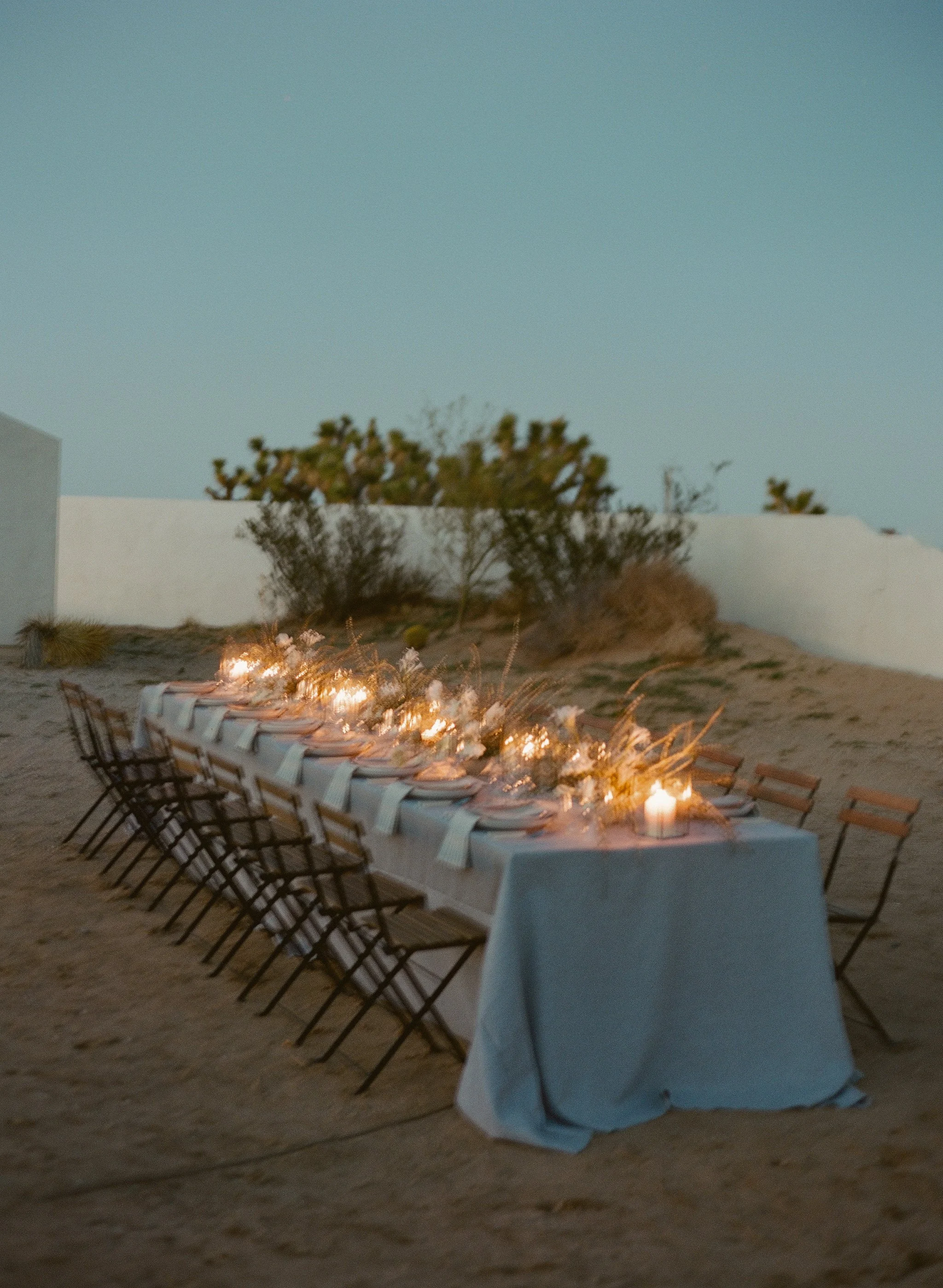 candlelit dinner table with a pale blue linen at dusk in the desert, joshua trees behind a white wall, on film