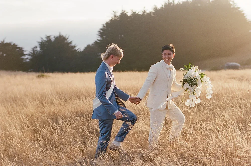 two grooms laughing and running through golden grass field at golden hour, big sur california — LGBTQ+ wedding film photography by Radostina Boseva
