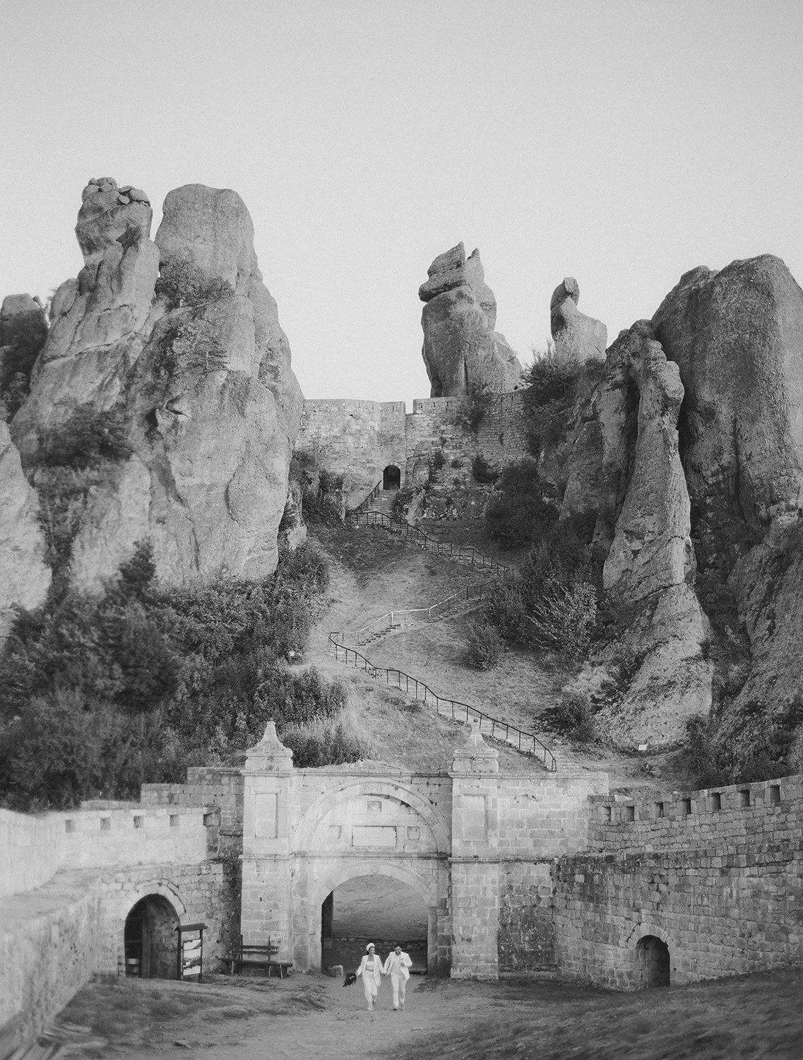 two figures in white walking through the fortress archway at belogradchik rocks · bulgaria destination elopement · documentary film photography by radostina boseva