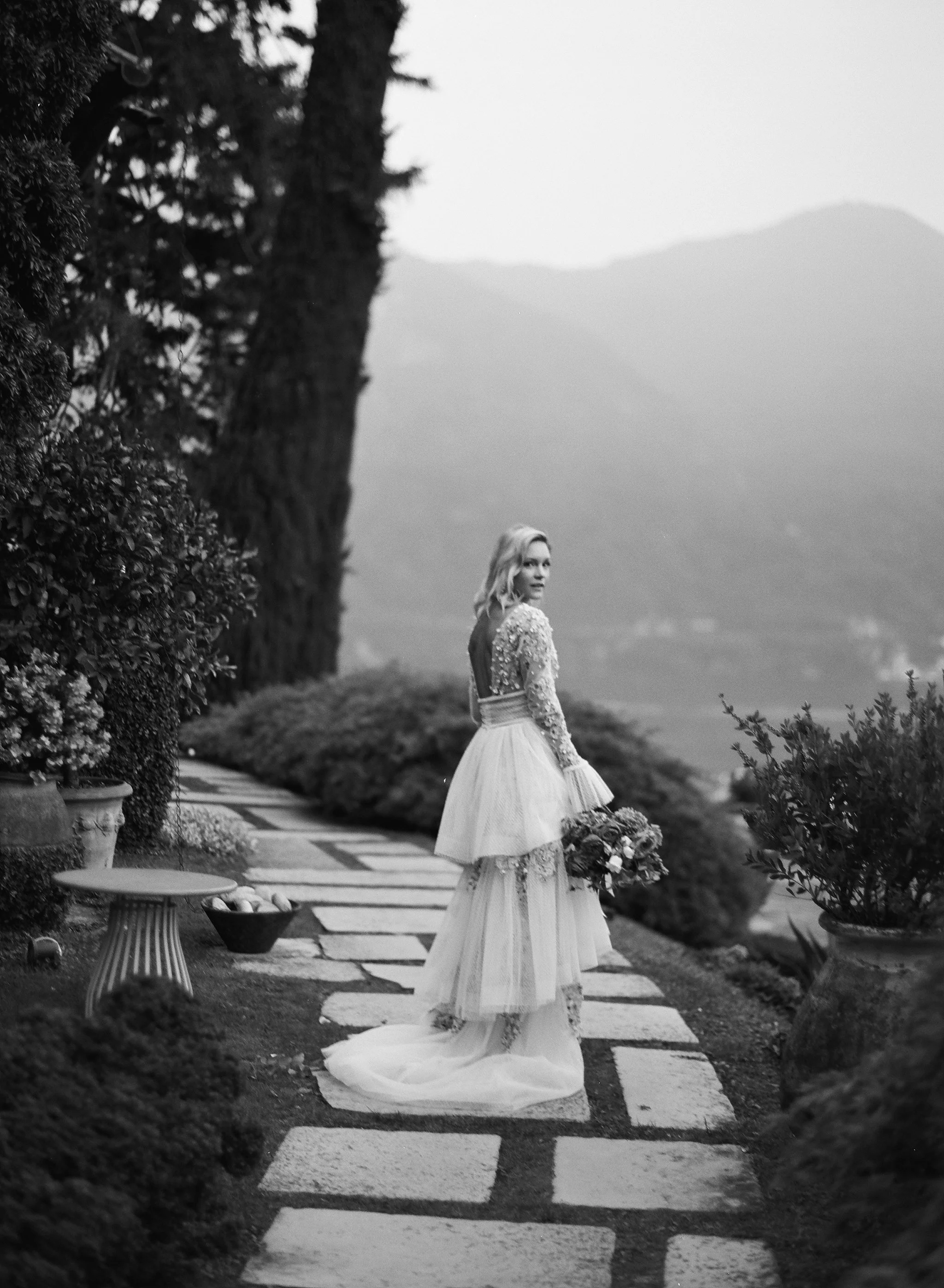 bride in a beaded long-sleeve gown with a tiered tulle skirt, looking back along a garden path · lake como elopement · black and white editorial film photography by radostina boseva