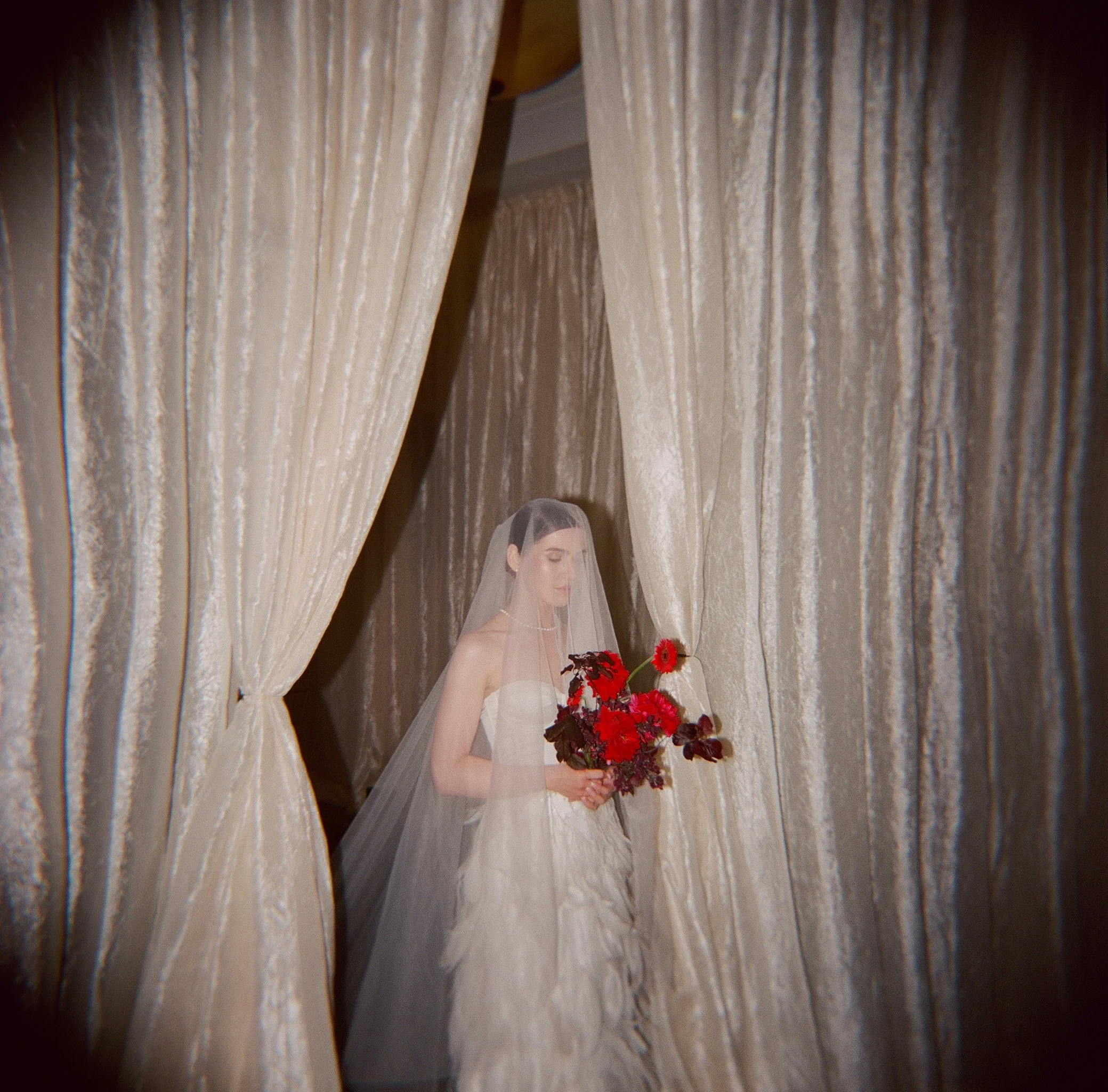 Bride in veil with red bouquet framed by dramatic curtains — film wedding photography, Los Angeles